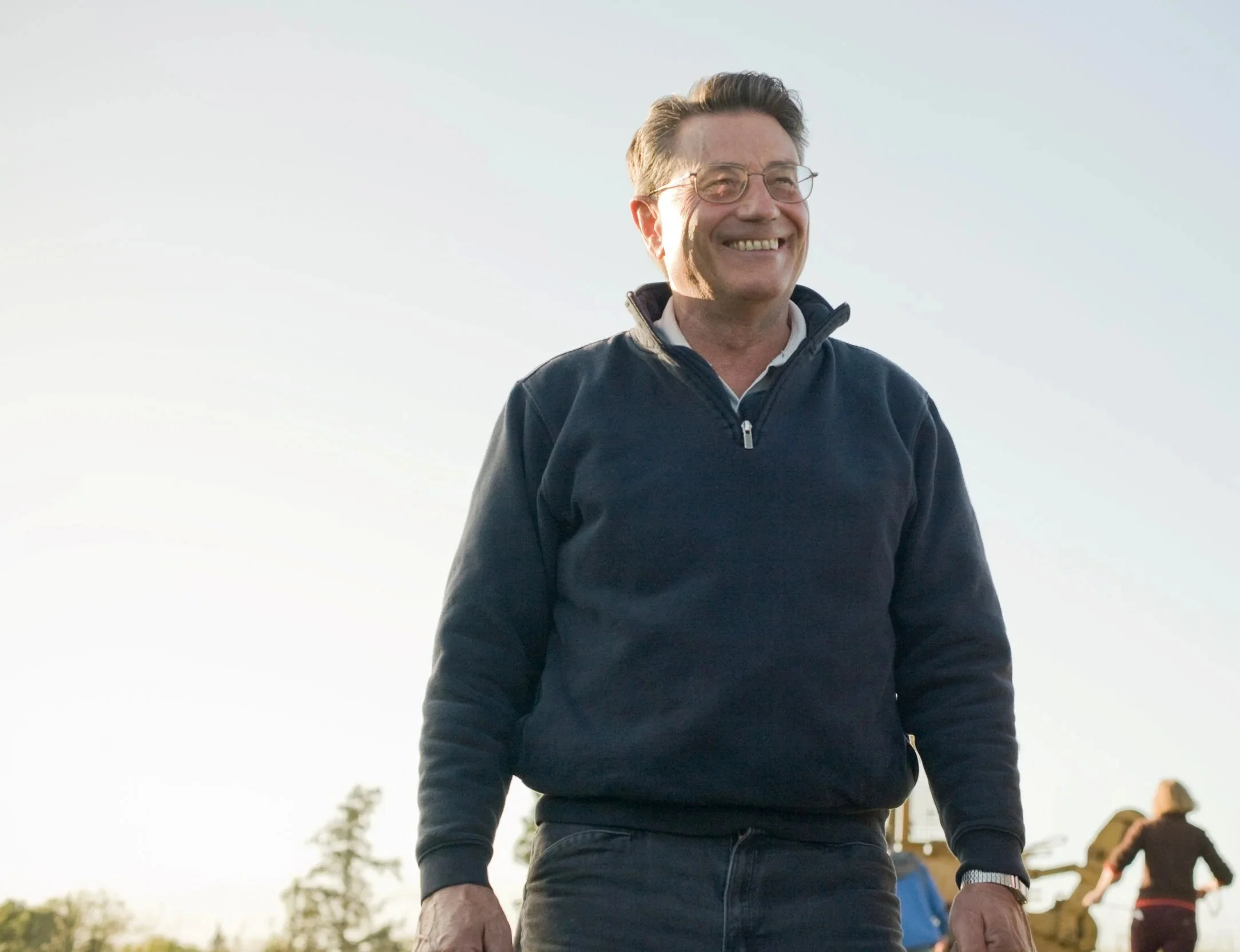 Smiling man wearing glasses and a navy blue zip-up jacket, standing outdoors under a clear sky.