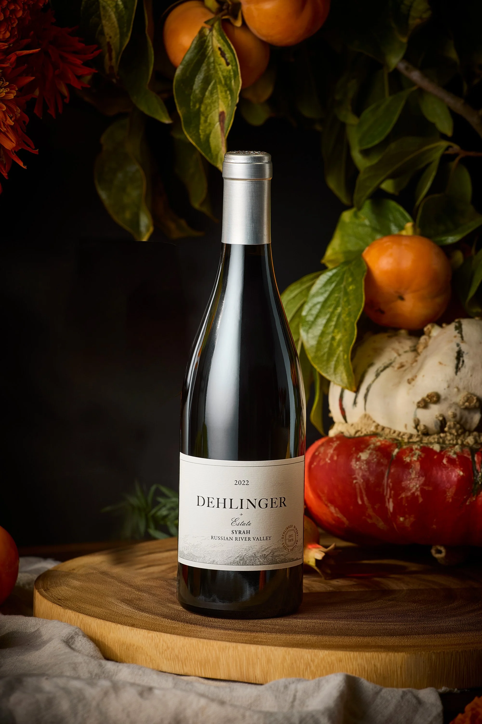 A bottle of Dehlinger Estate Syrah wine from 2022 placed on a wooden surface. The background features various fruits and leaves, including apples, pomegranates, and gourds, against a dark setting.