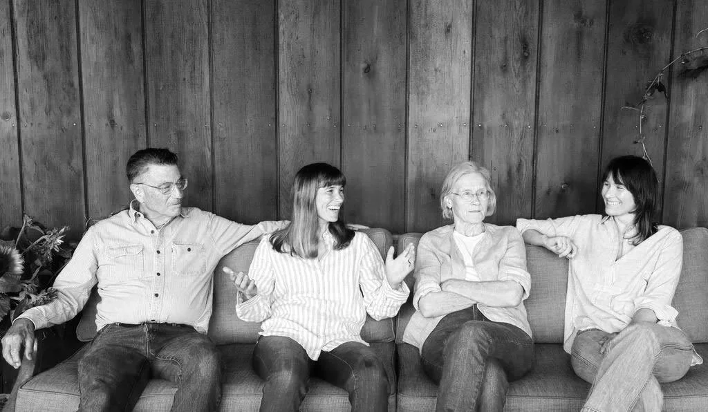Four people are sitting on a couch against a wooden wall, engaging in conversation and smiling. The group consists of two women and two men, with one woman and one man appearing older.