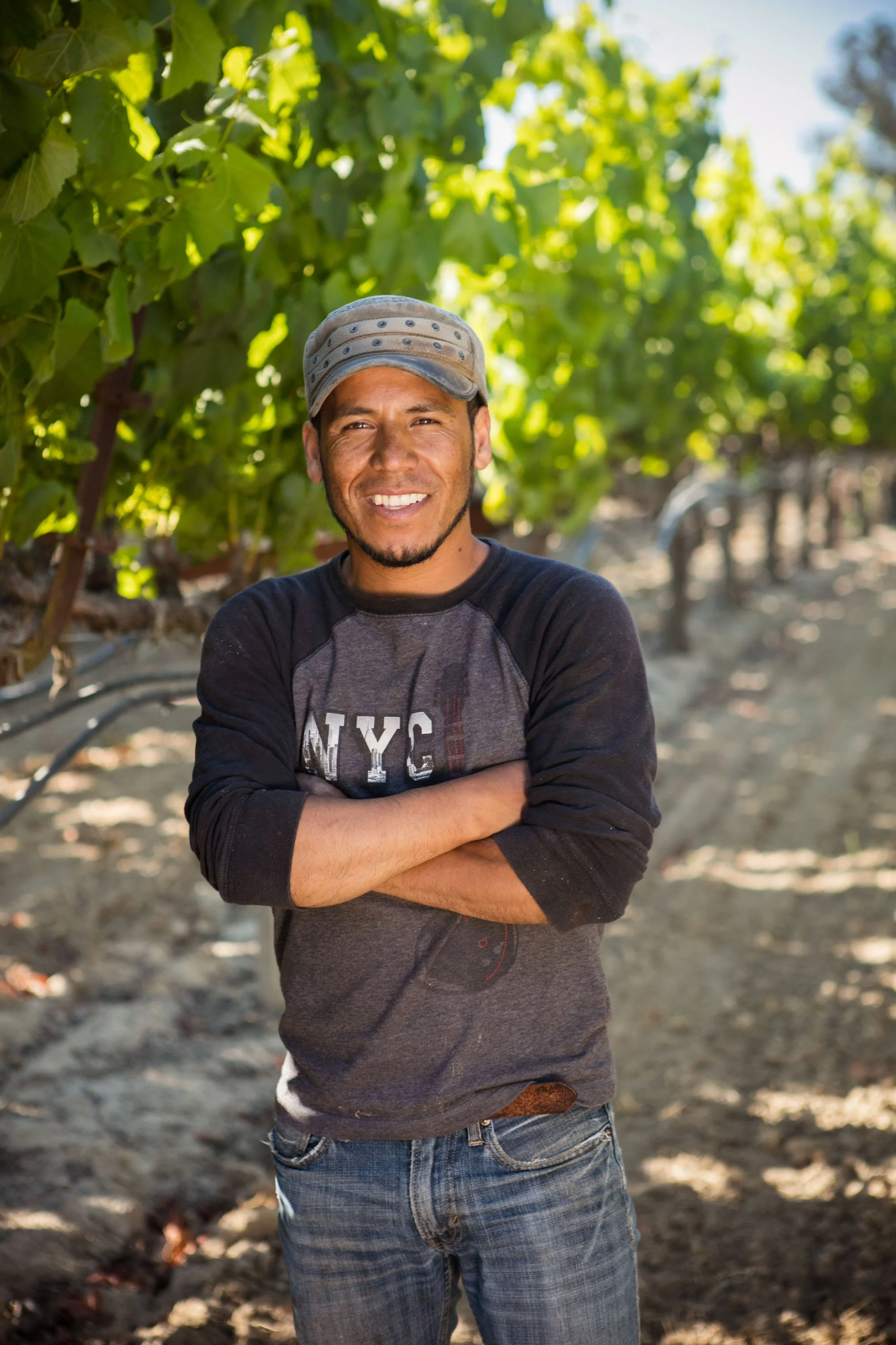 A man stands outdoors in front of green leaves, smiling with his arms crossed, wearing a baseball cap, a dark long-sleeve shirt, and jeans.