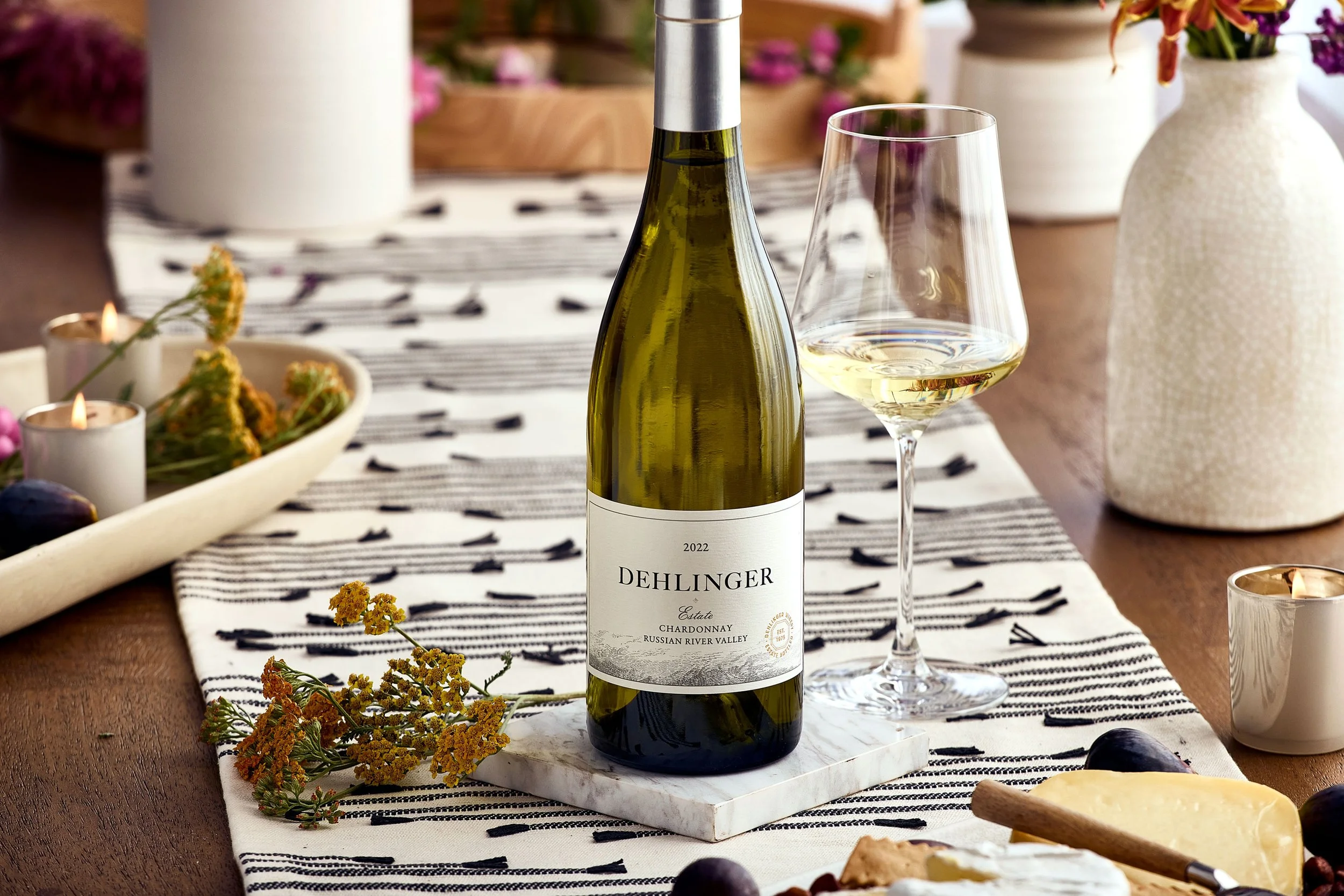 A bottle of Dehlinger estate Chardonnay wine with a glass of white wine beside it on a table with a black and white striped table runner, surrounded by flowers, candles, cheese, and grapes.