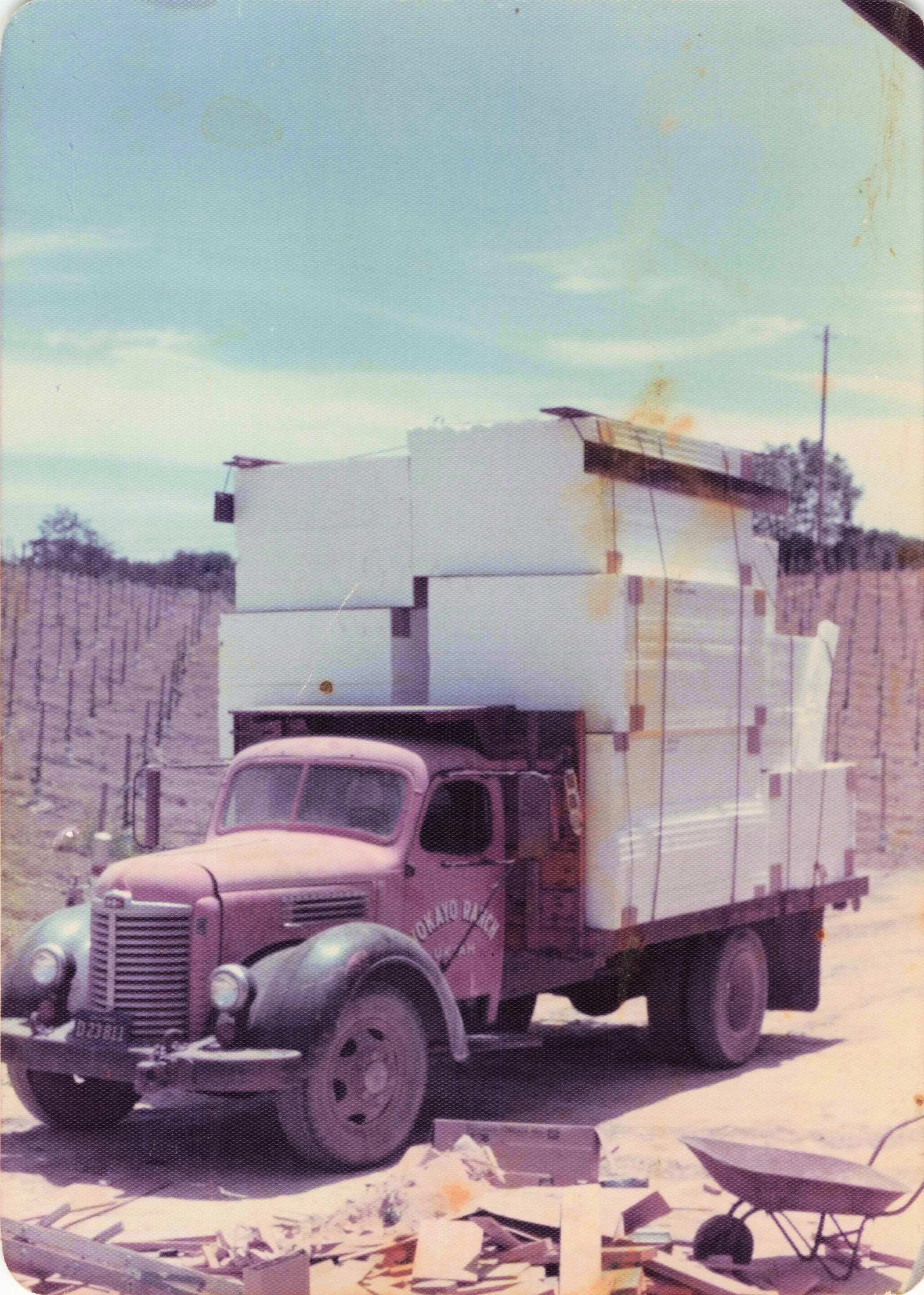Vintage truck with a cargo of large white boxes, parked outdoors with debris including wood and metal scattered on the ground.
