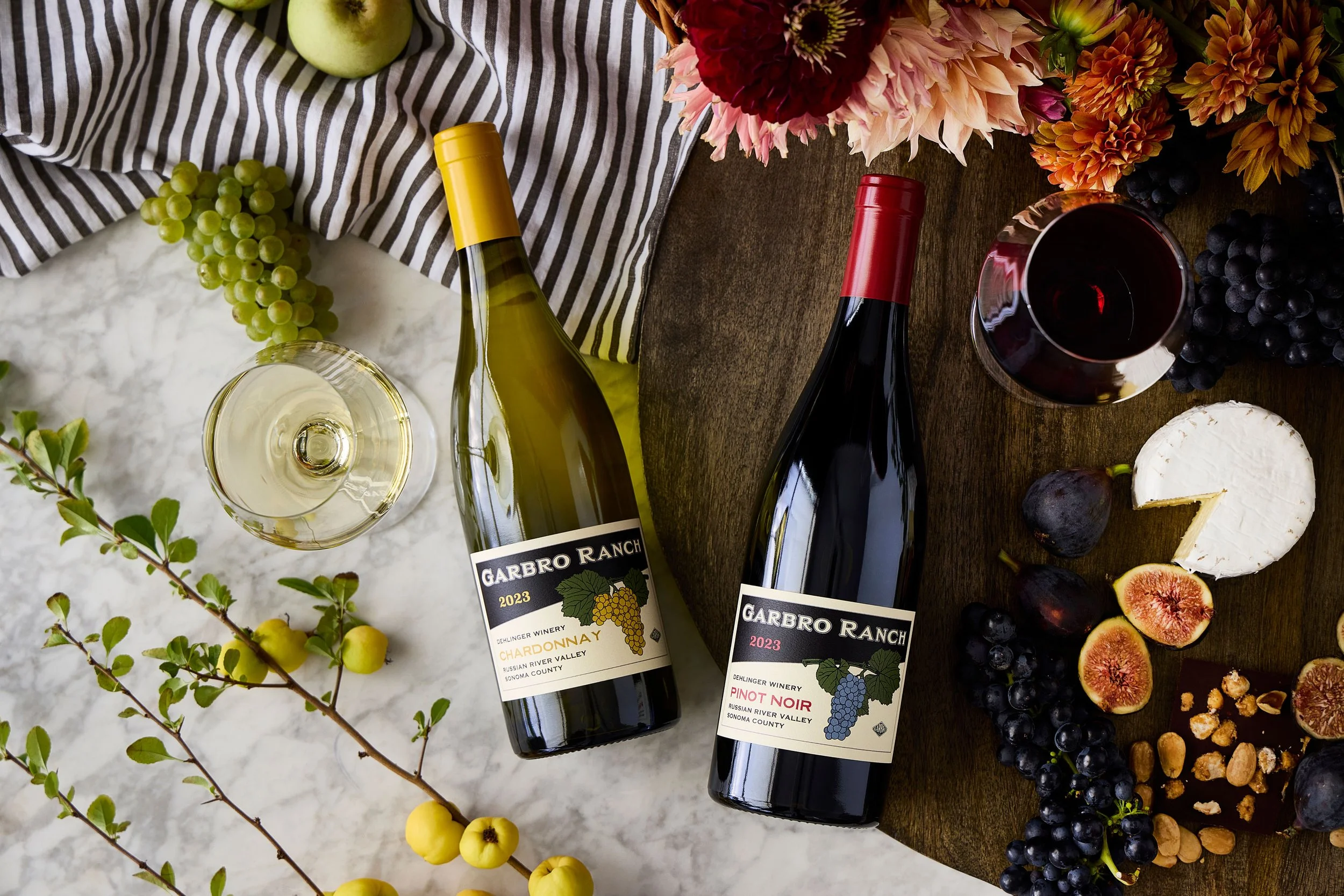 Two bottles of red wine, one labeled 'Garbro Ranch 2023 Chardonnay' and the other 'Garbro Ranch 2023 Pinot Noir', are surrounded by grapes, figs, cheese, honey, and abundant colorful flowers on a wooden table.