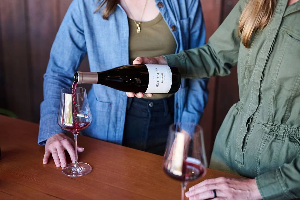 A woman pours a bottle of Dehlinger wine for another person. The wine glass is resting on a wooden surface