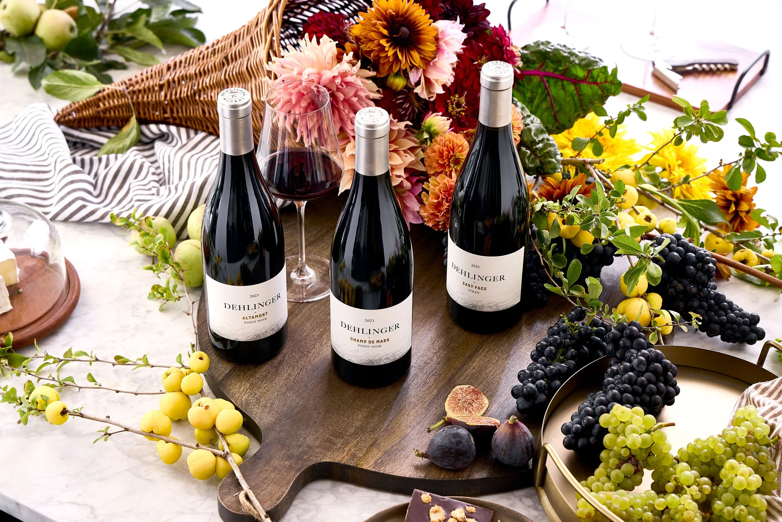 Three bottles of Dehlinger wine, a filled wine glass, and a variety of colorful flowers and fruits arranged on a table for a celebration or gathering.