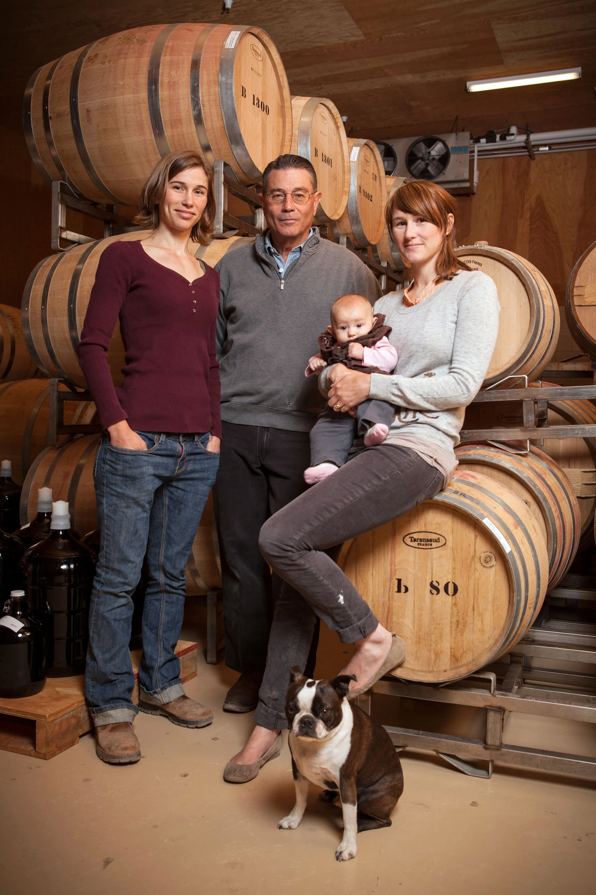 Four people, two women, a man, and a baby, standing in a winery cellar with large wooden barrels. A dog is sitting in front of the woman on the right.
