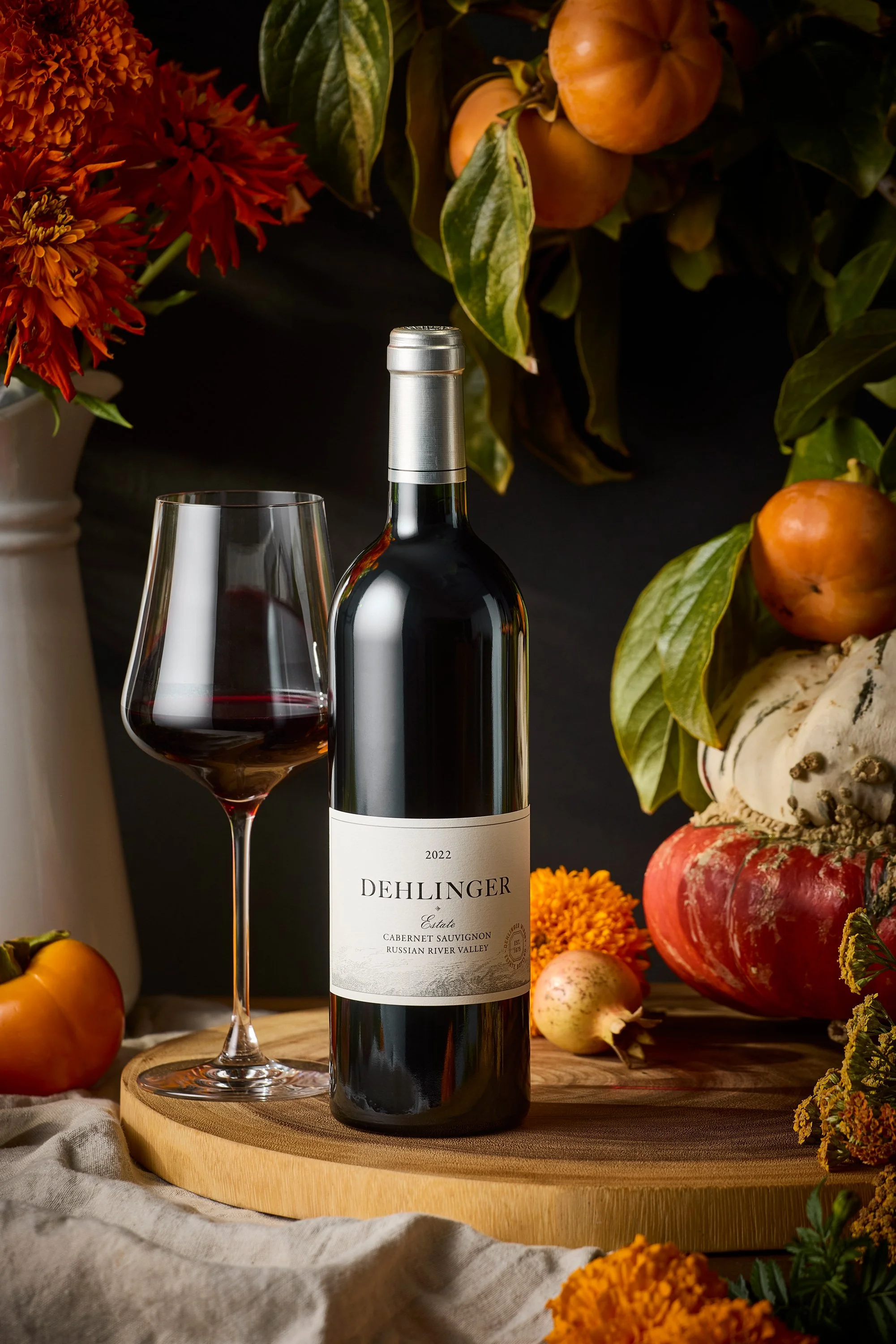 A bottle of Dehlinger 2022 estate Cabernet Sauvignon wine with a glass of red wine beside it. The scene includes autumnal flowers and vegetables such as pumpkins, gourds, and marigolds, set on a wooden tray and table with a dark background.