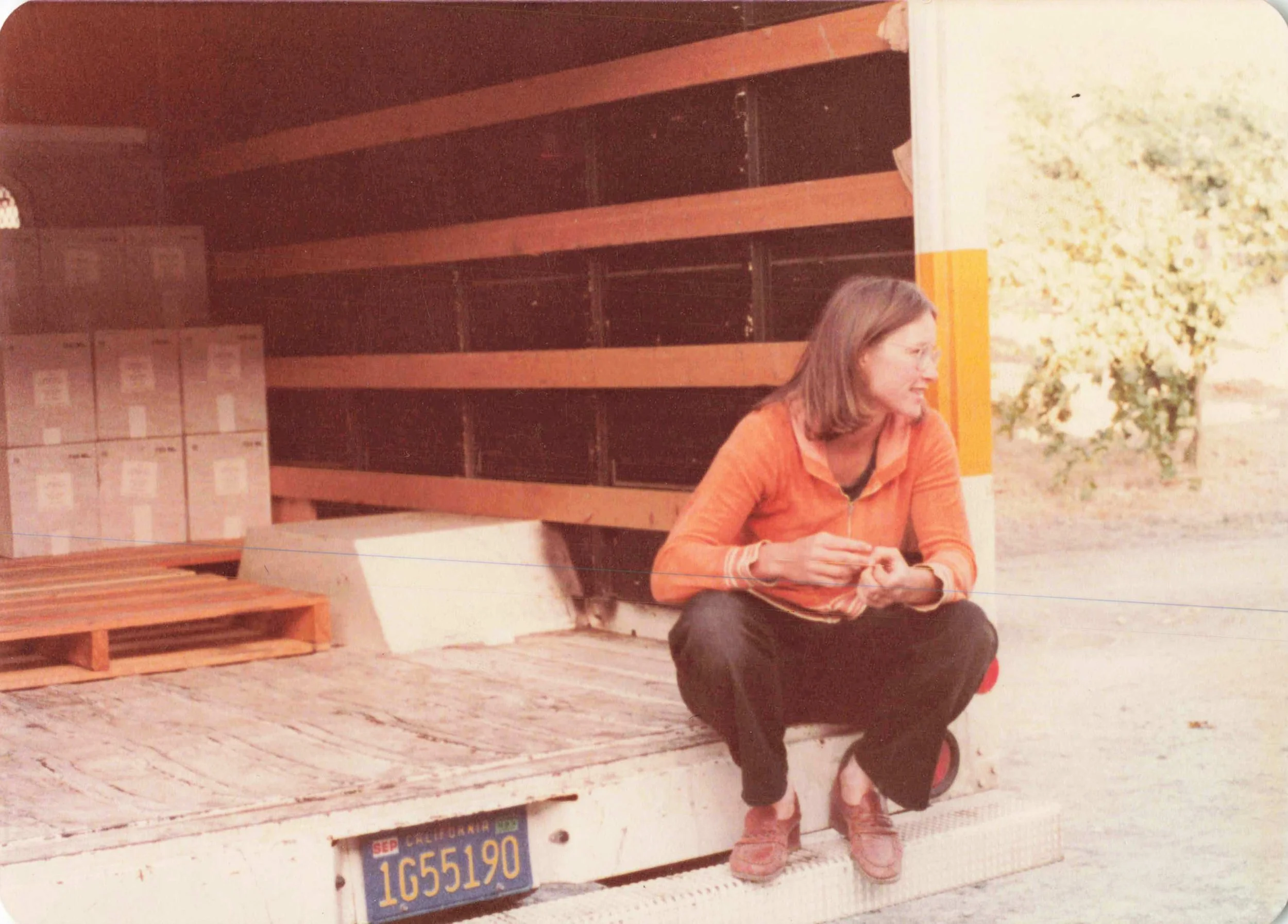 A woman with long hair, glasses, and a pink jacket sitting on the back of a pickup truck with a woman with long dark hair sitting cross-legged nearby. The truck has a California license plate, and there are stacks of boxes and a wooden pallet in the background.