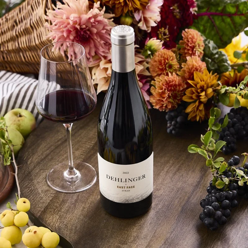 A bottle of Dehlinger East Face Syrah wine, a glass of red wine, and grapes on a wooden table surrounded by colorful flowers and greenery.
