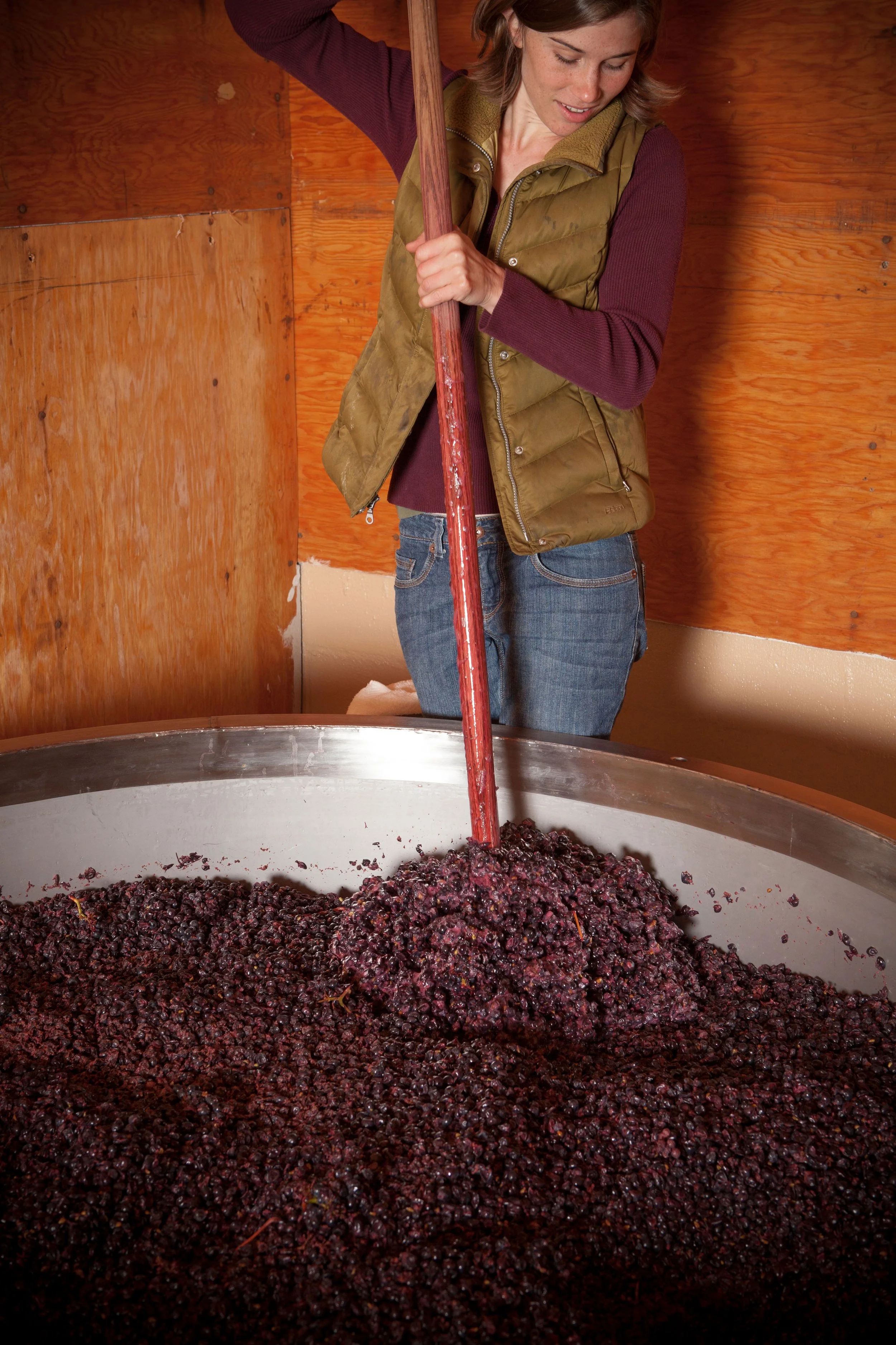 A woman stirring grape mash in a large metal fermentation vat during winemaking, with wooden walls in the background.