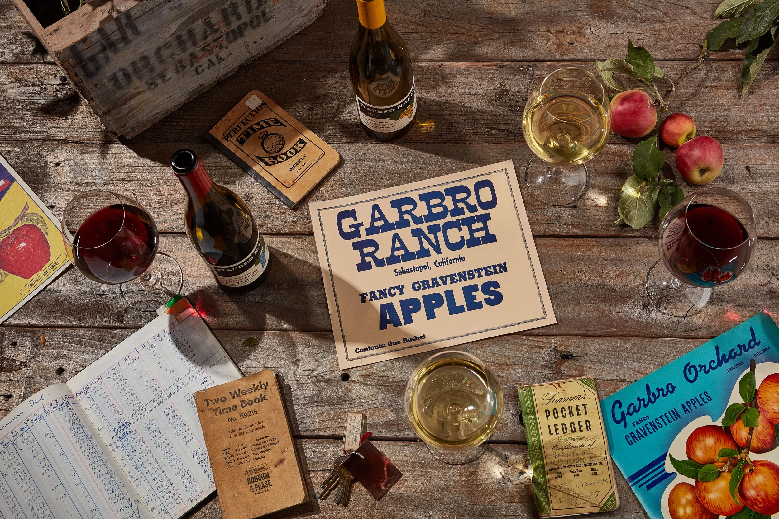 A rustic wooden table with a bottle of Garbro Ranch wine, two glasses of white wine, red apples with green leaves on a branch, a vintage pocket ledger book, a farmers pocket book, a GARDBRO RANCH sign, a handwritten notebook with a pen, and a blue pamphlet about Garbro Ranch Apples.