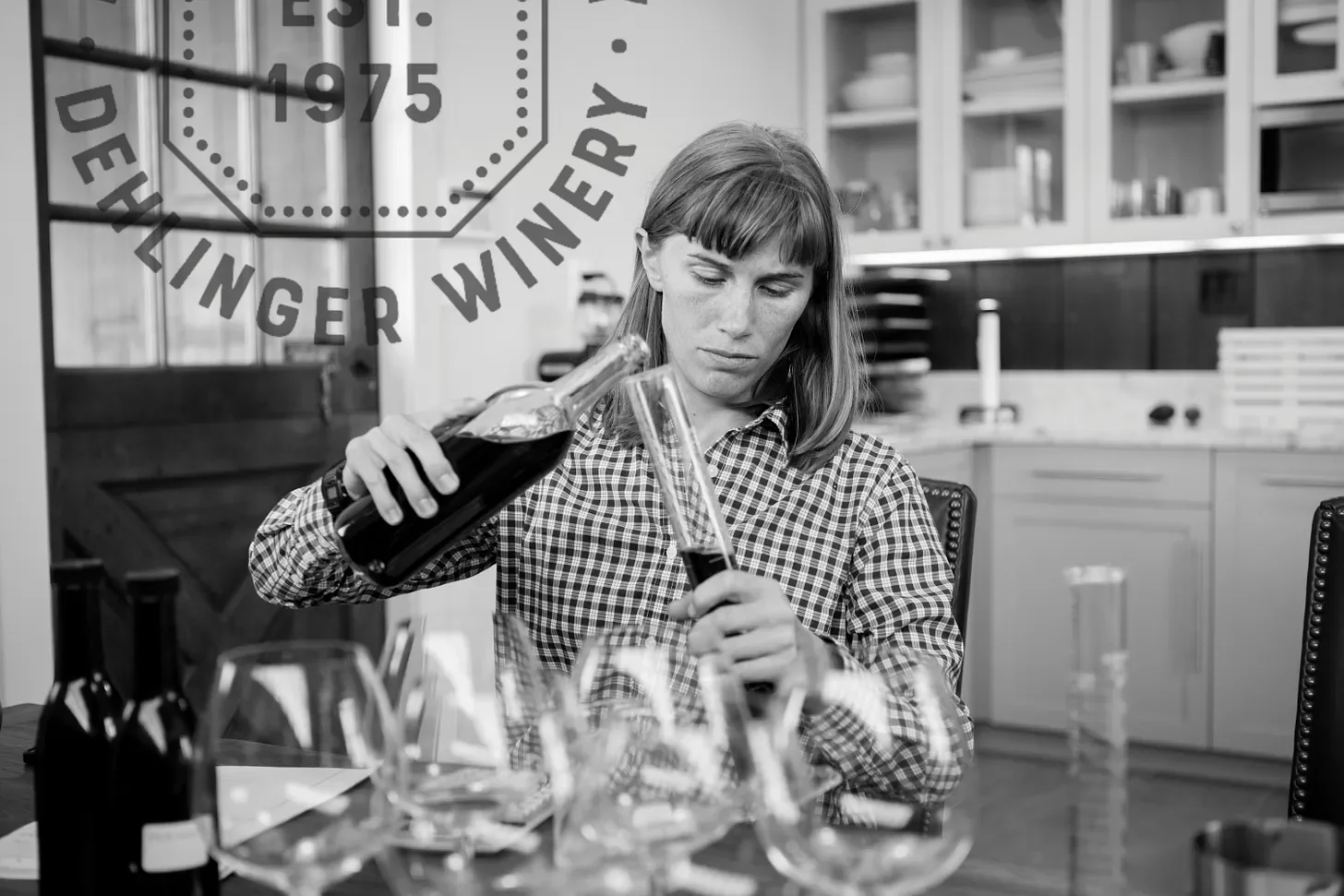 A woman with straight hair and a checkered shirt pouring wine from a bottle into a glass at a dining table, with other glasses and bottles in front of her. In the background, there is a kitchen with cabinets and shelves. The image is in black and white.