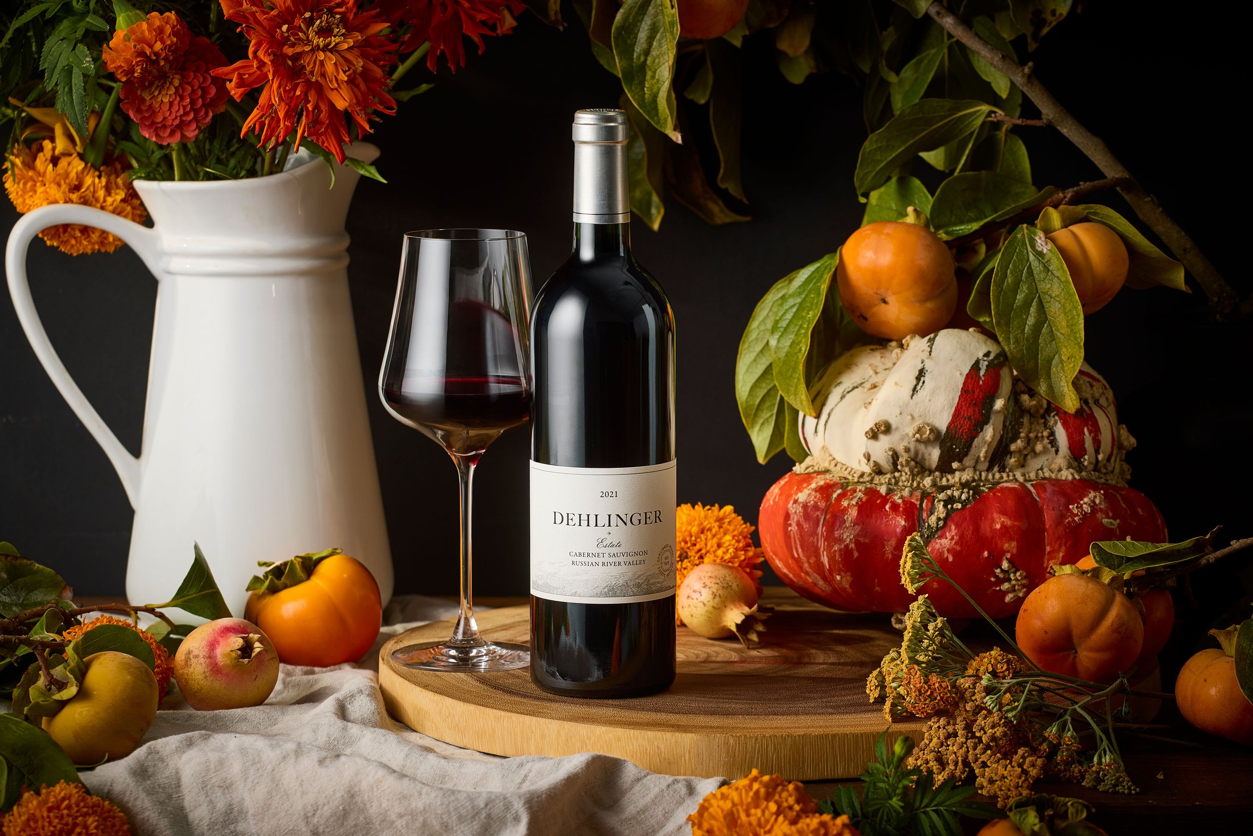 A bottle of Dehlinger Estate Cabernet Sauvignon red wine, a glass filled with red wine, a white vase with orange and red flowers, and various fruits and flowers arranged on a wooden surface and surrounded by green leaves and flowers.