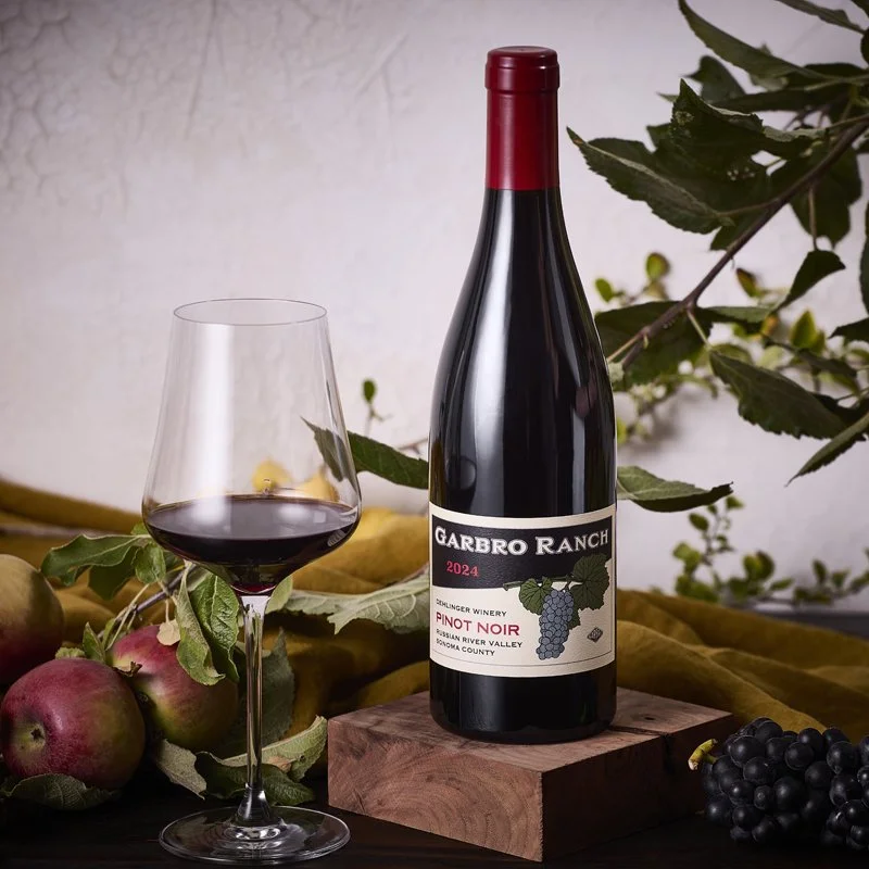 A bottle of Garbro Ranch Pinot Noir 2024 wine on a wooden block, with a filled wine glass beside it, surrounded by apples, grapes, and grapevine leaves.
