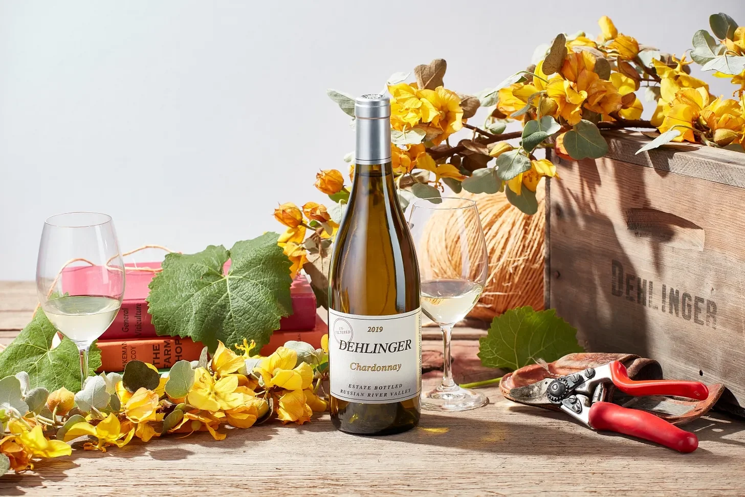 A bottle of Dehlinger Chardonnay wine on a wooden table with two glasses of white wine, yellow flowers, green leaves, a pair of pruning shears, and a wooden crate labeled Dehlinger in the background.