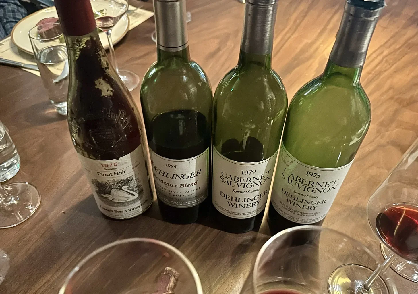 Four wine bottles on a wooden table, with glasses of red and white wine surrounding them. The bottles are corked and labeled with different years and winery names.