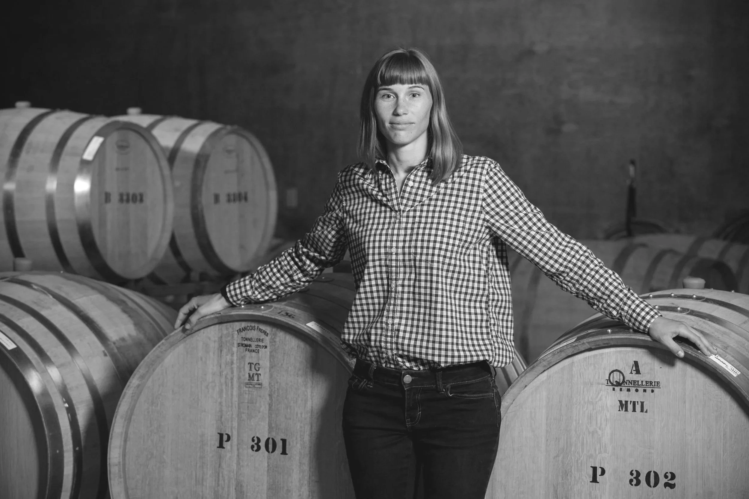 A woman standing in a winery, touching large wooden wine barrels with her right hand and arms akimbo, wearing a checked shirt and dark jeans, with a neutral expression on her face.