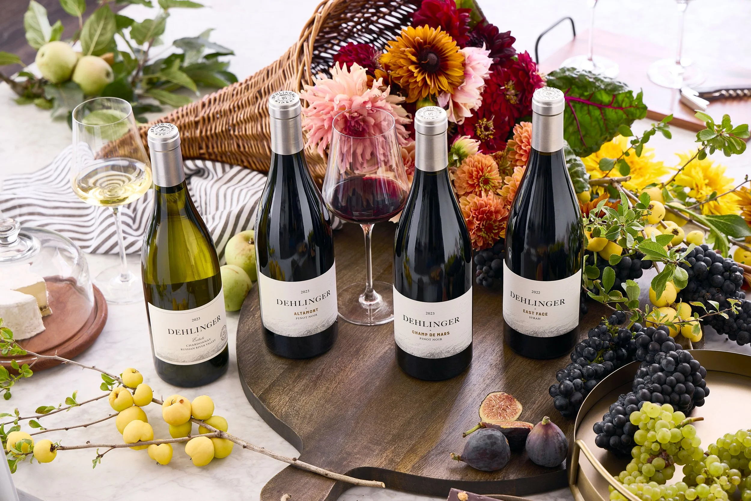 A table with five bottles of Dehlinger wine, two glasses of white and red wine, a floral arrangement, grapes, apples, figs, and a cheese platter, surrounded by greenery and flowers.