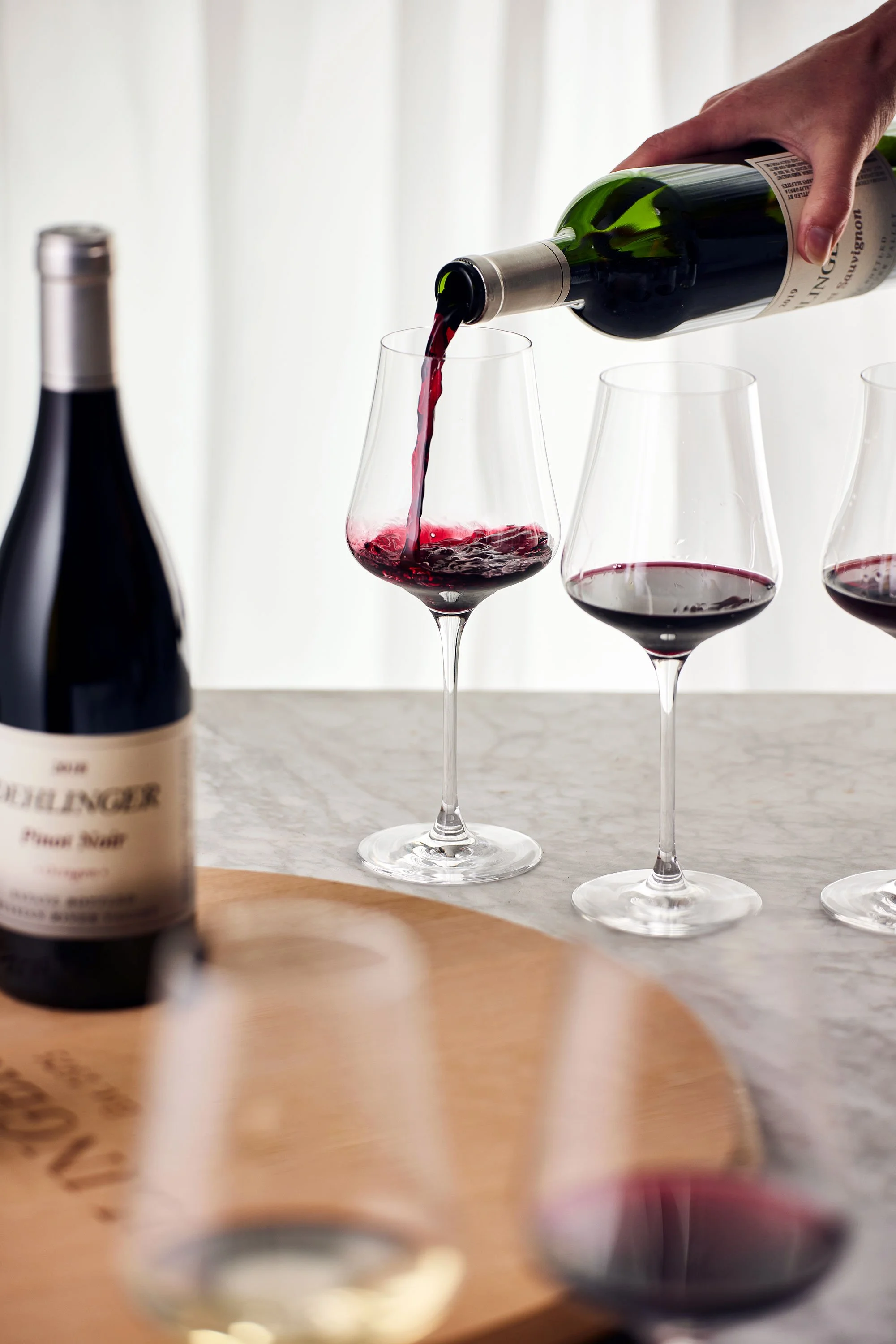 Hand pouring red wine into a wine glass, with additional wine glasses and a wine bottle on a wooden table.