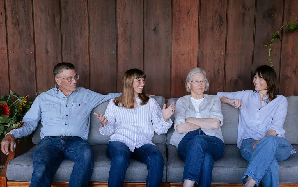 Four adults sitting on a gray couch, engaging in conversation with a wooden wall background. The group includes an elderly woman with glasses, a middle-aged woman, an older man, and a young woman, all smiling or talking.