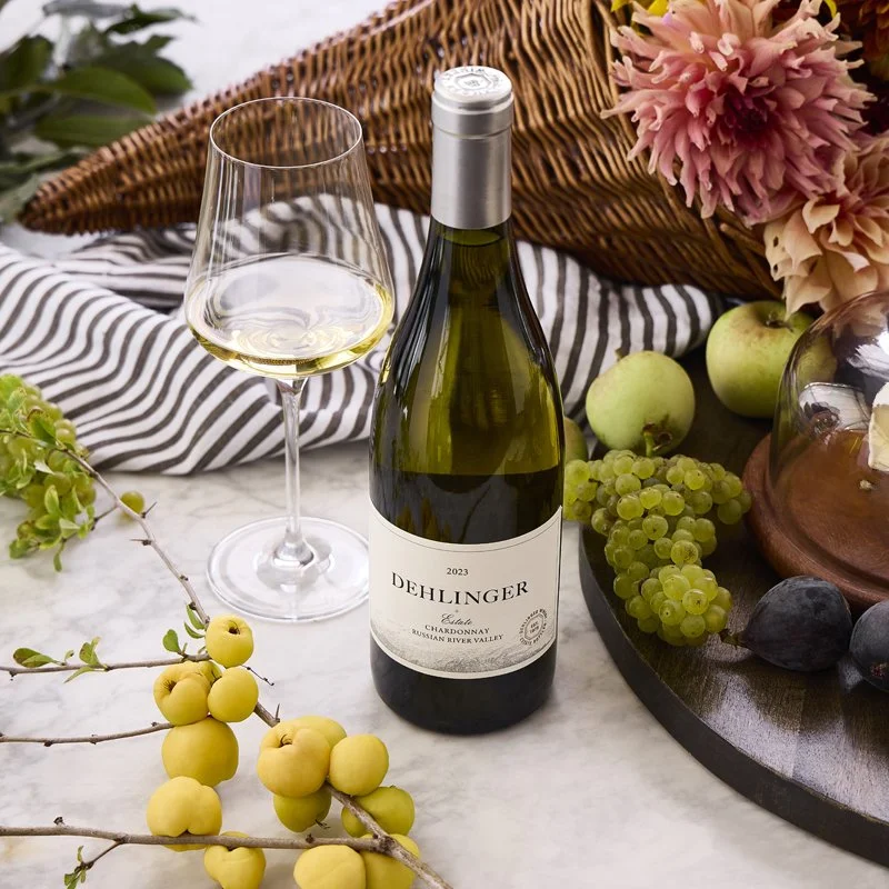 A bottle of Dehlinger Chardonnay wine, a filled wine glass, green and purple grapes, green apples, and yellow berries on a table decorated with flowers and a wicker basket.