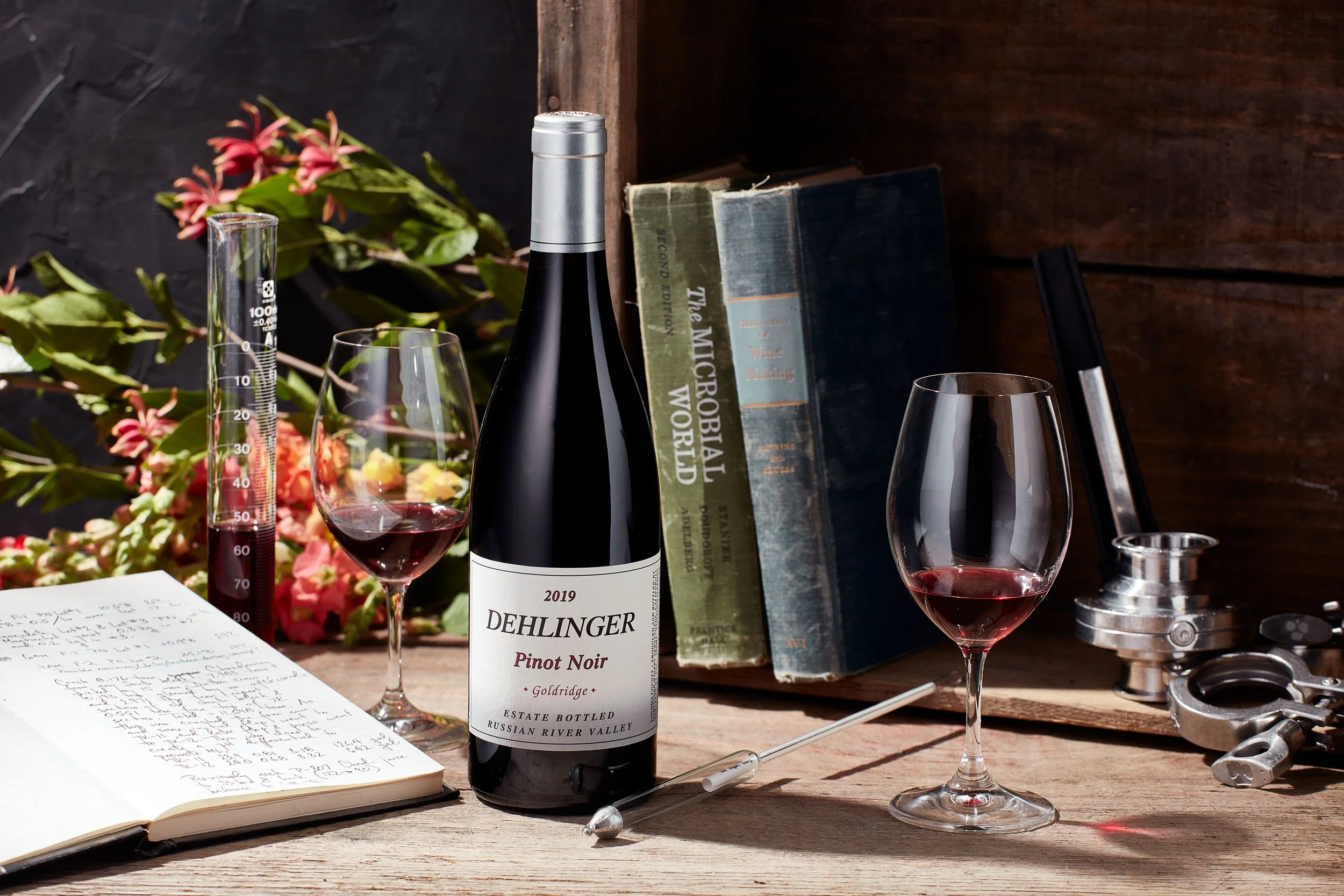 A wine bottle labeled Dehlinger Pinot Noir from 2019, two glasses of red wine, a notebook with handwritten notes, an open book titled "The Microbial World," a pen, a syringing tool, a pair of scissors, a flower arrangement, a test tube, and a wine stopper on a wooden surface, with dark background and some flowers.