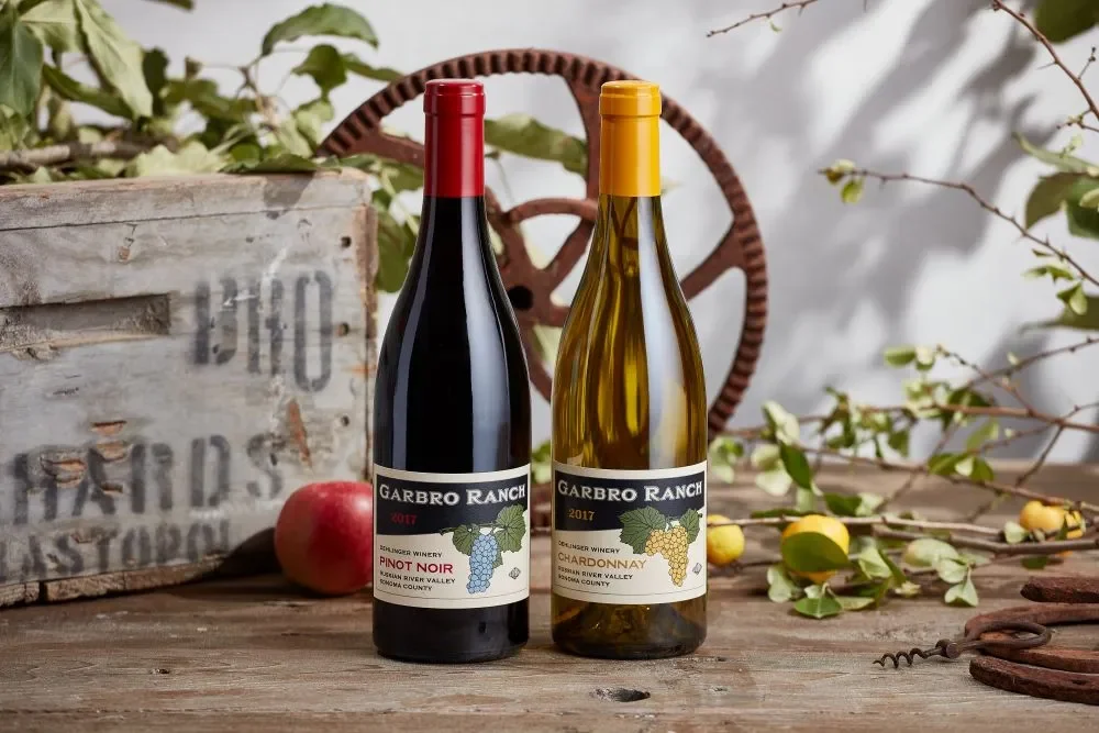 Two wine bottles, one Pinot Noir and one Chardonnay, on a rustic wooden surface with a wooden box, apple, and branches with yellow fruit and green leaves in the background.