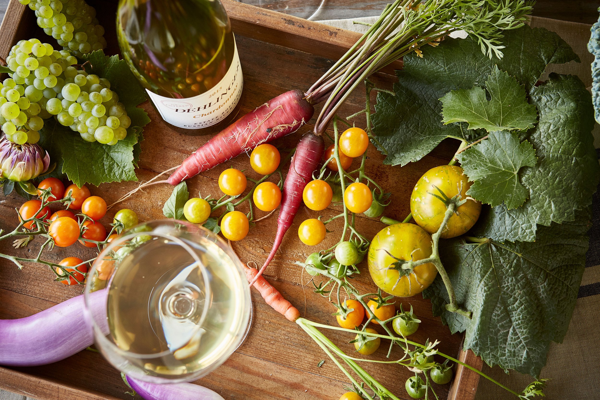 A wooden tray holding fresh vegetables, a bottle of white wine, and a glass of wine. The vegetables include pinkish-red radishes with green leaves, yellow cherry tomatoes, red cherry tomatoes, an eggplant, purple carrots, and dark green leafy greens.