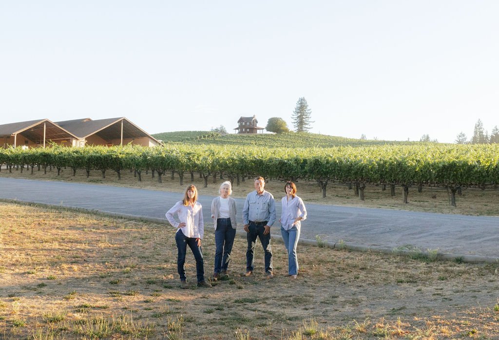 Four people standing on a vineyard landscape with hills, a house, and trees in the background.