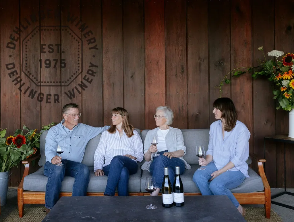 Four people sitting on a gray couch inside a winery, holding glasses of wine, with flowers on each side and bottles on a table in front of them.
