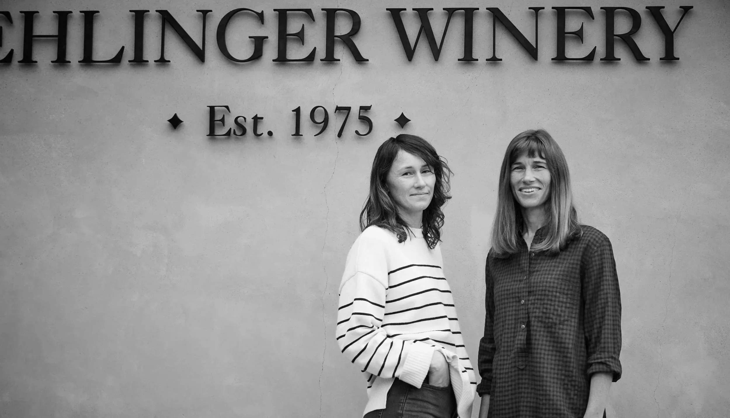 Two women standing in front of a wall with a sign that reads 'Chilinger Winery Est. 1975'. The woman on the left has wavy hair, wearing a striped sweater, and the woman on the right has straight hair with bangs, wearing a checkered shirt.