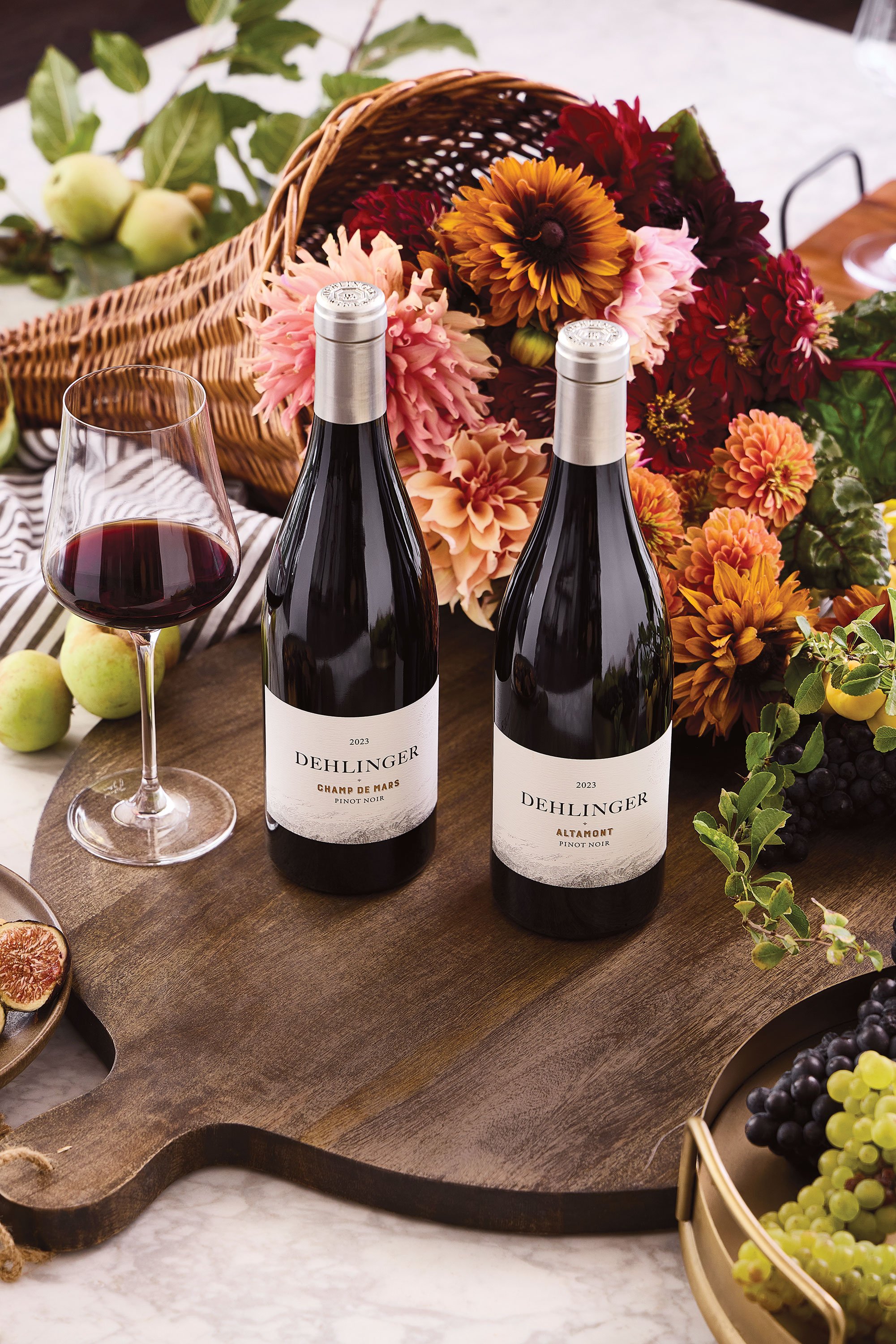 Two bottles of Dehlinger Pinot Noir wine next to a glass of red wine on a wooden tray, with a basket of colorful flowers and green apples in the background.