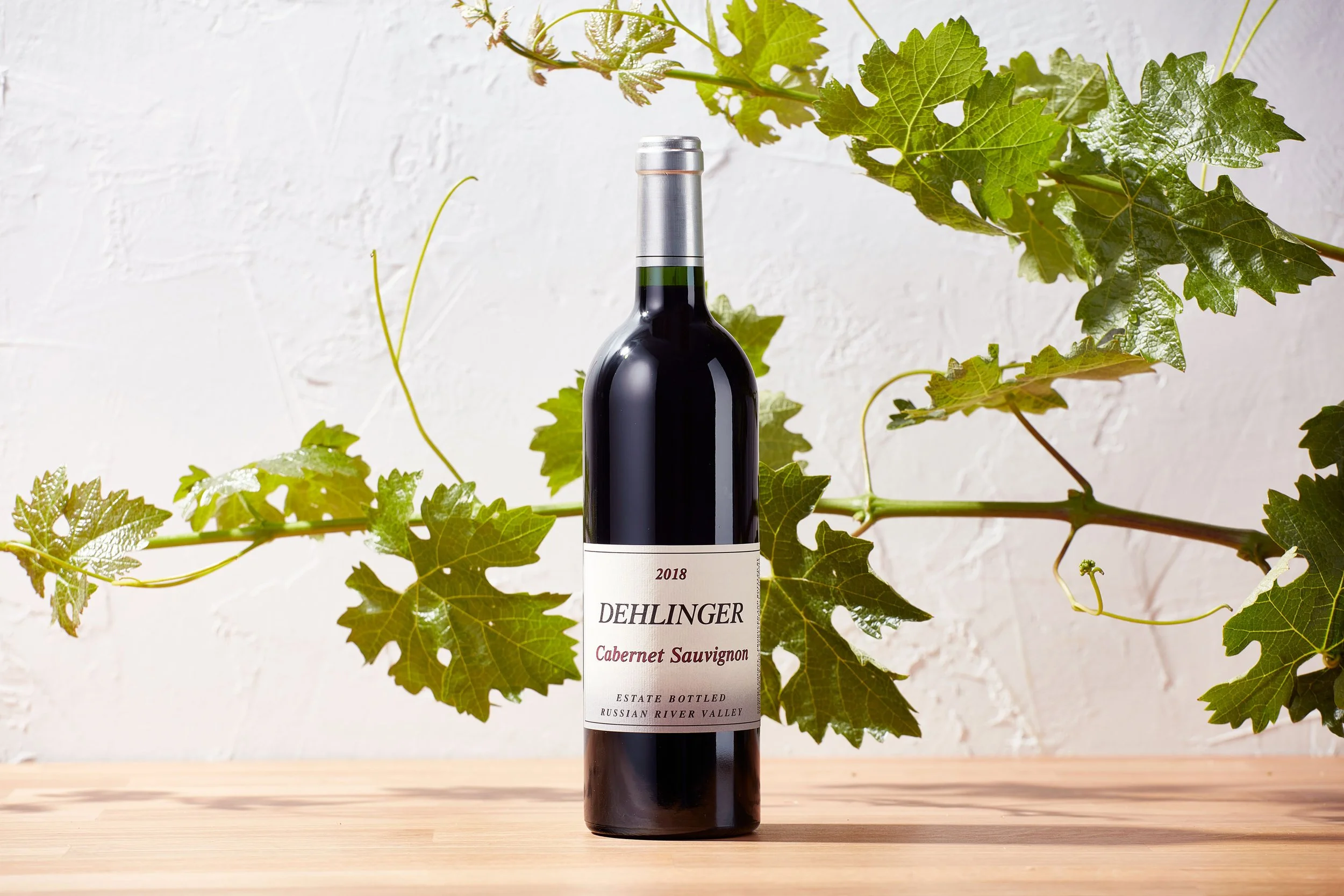 A bottle of red wine labeled Dehlinger Cabernet Sauvignon 2018, placed on a wooden surface with green grapevine leaves in the background