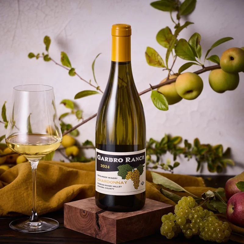 A bottle of Garbro Ranch Chardonnay wine, vintage 2024, from Alexander Valley, Sonoma County, sits on a wooden block, with a glass of white wine beside it, surrounded by grapes, apples, and a branch with green apples, against a neutral background.