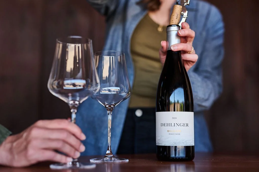 A hand opens a bottle of Dehlinger wine while a second person's hand waits with expection