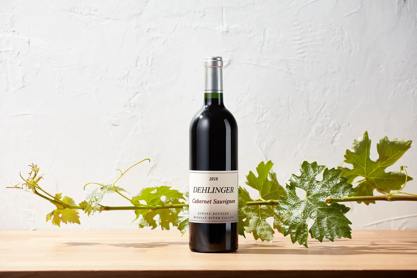 A bottle of Dehlinger Cabernet Sauvignon wine from 2018, estate bottled in Russian River Valley, placed on a wooden surface with green grapevine leaves behind it.