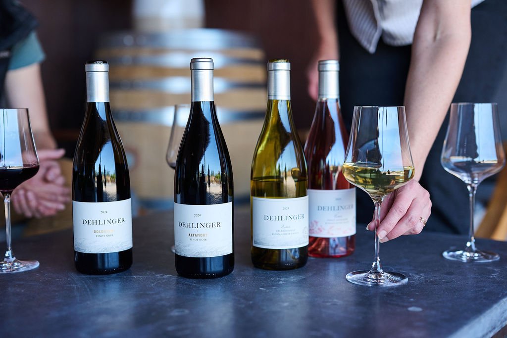 A hand placing a glass of Chardonnay in front of three bottles of Dehlinger wine