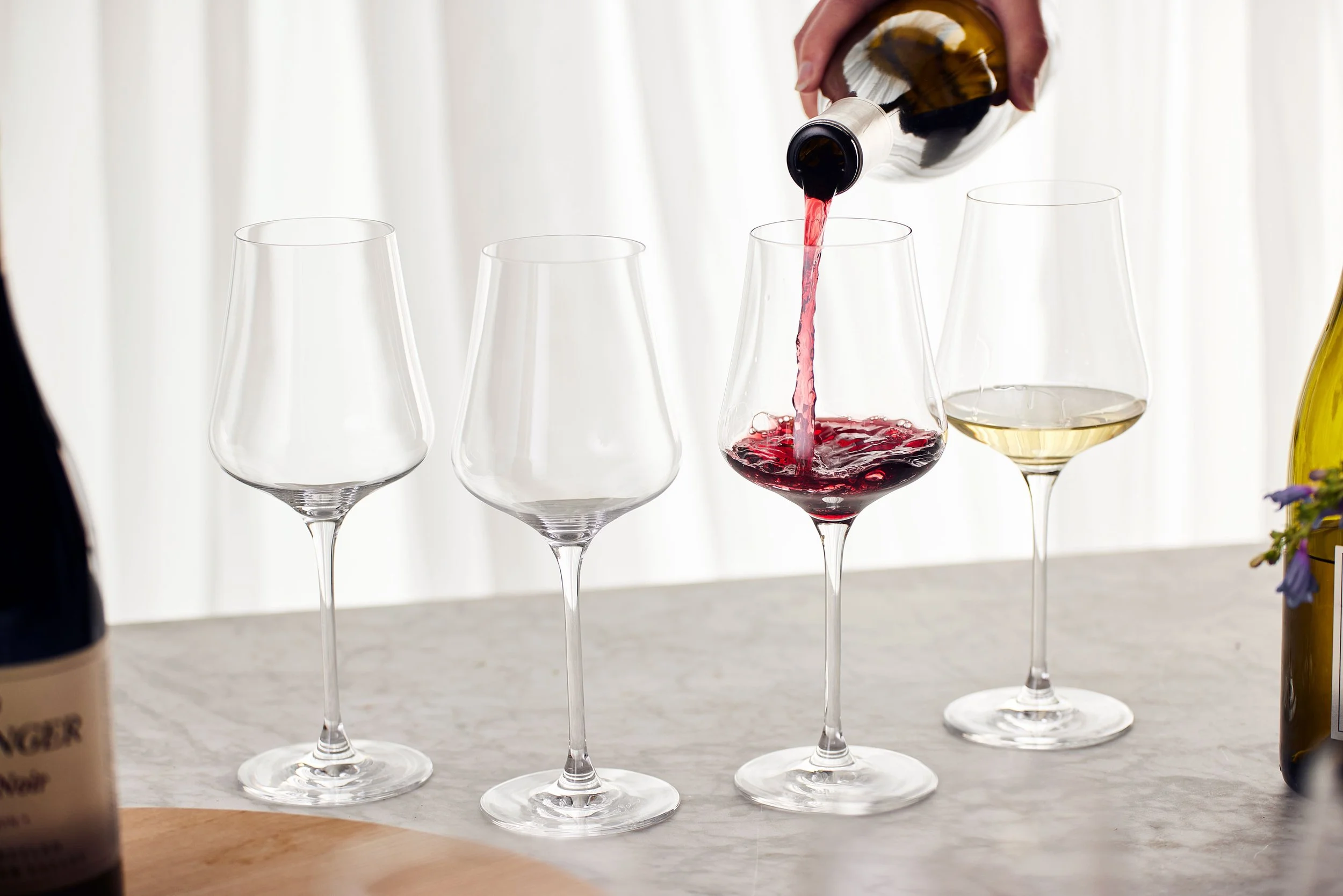 A hand pouring red wine into a stemmed wine glass, with empty wine glasses nearby on a light-colored surface, with a white curtain background.