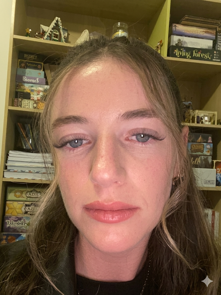 A young woman with light skin, blue eyes, and wavy light brown hair standing in front of a bookshelf filled with various books and board games.