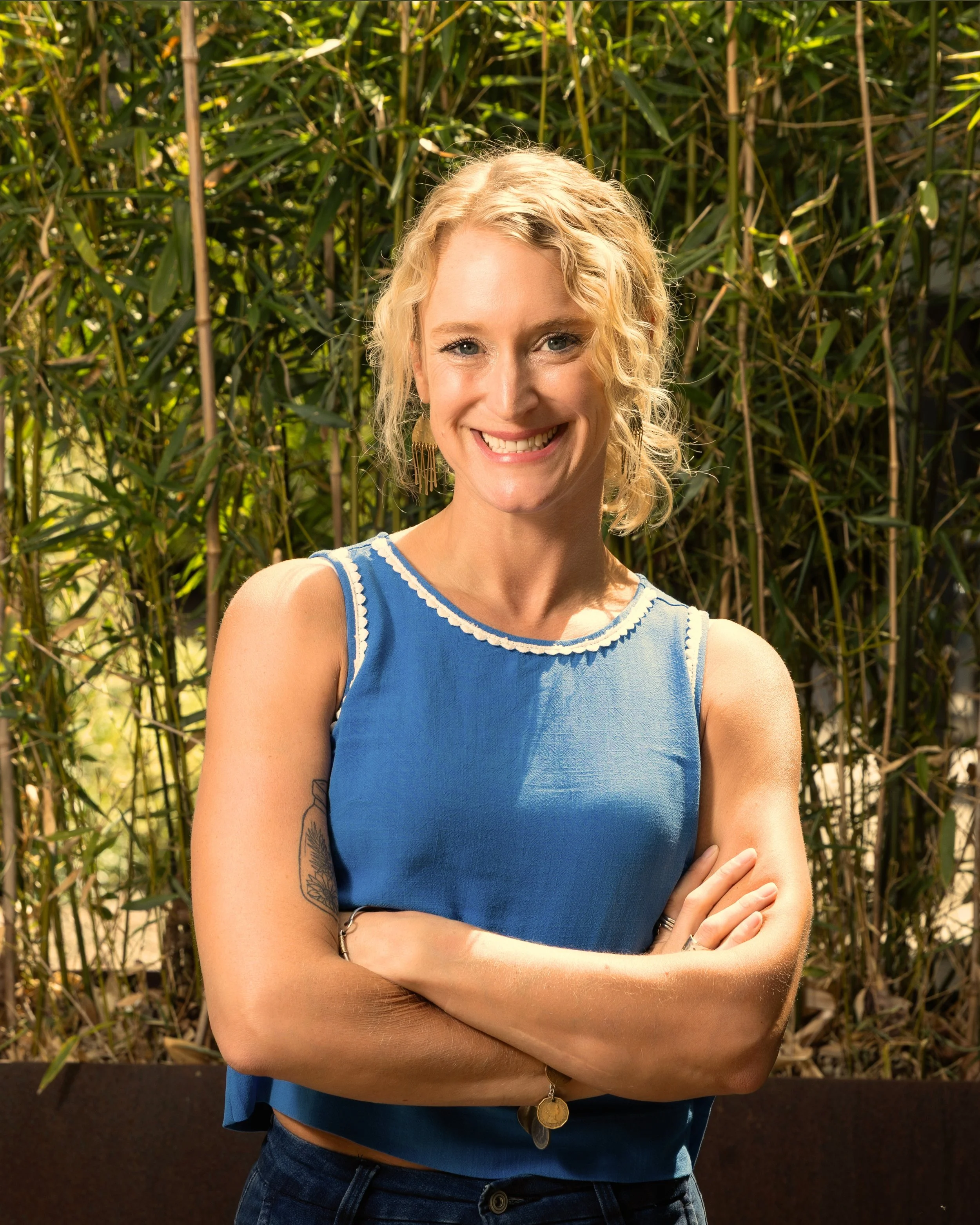 A woman with blonde curly hair smiling and crossing her arms in front of a backdrop of tall green grass.