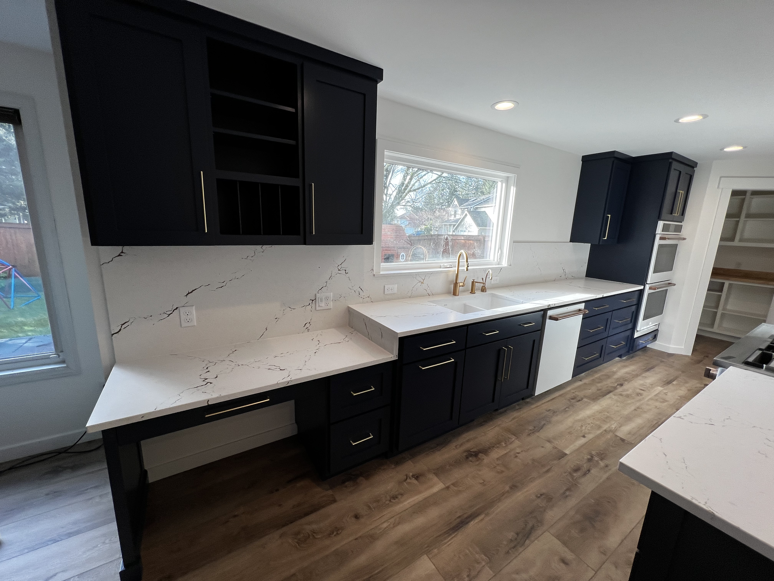 Modern kitchen with white marble countertops, black cabinetry with gold handles, a double oven, a large window above the sink, and wood flooring.