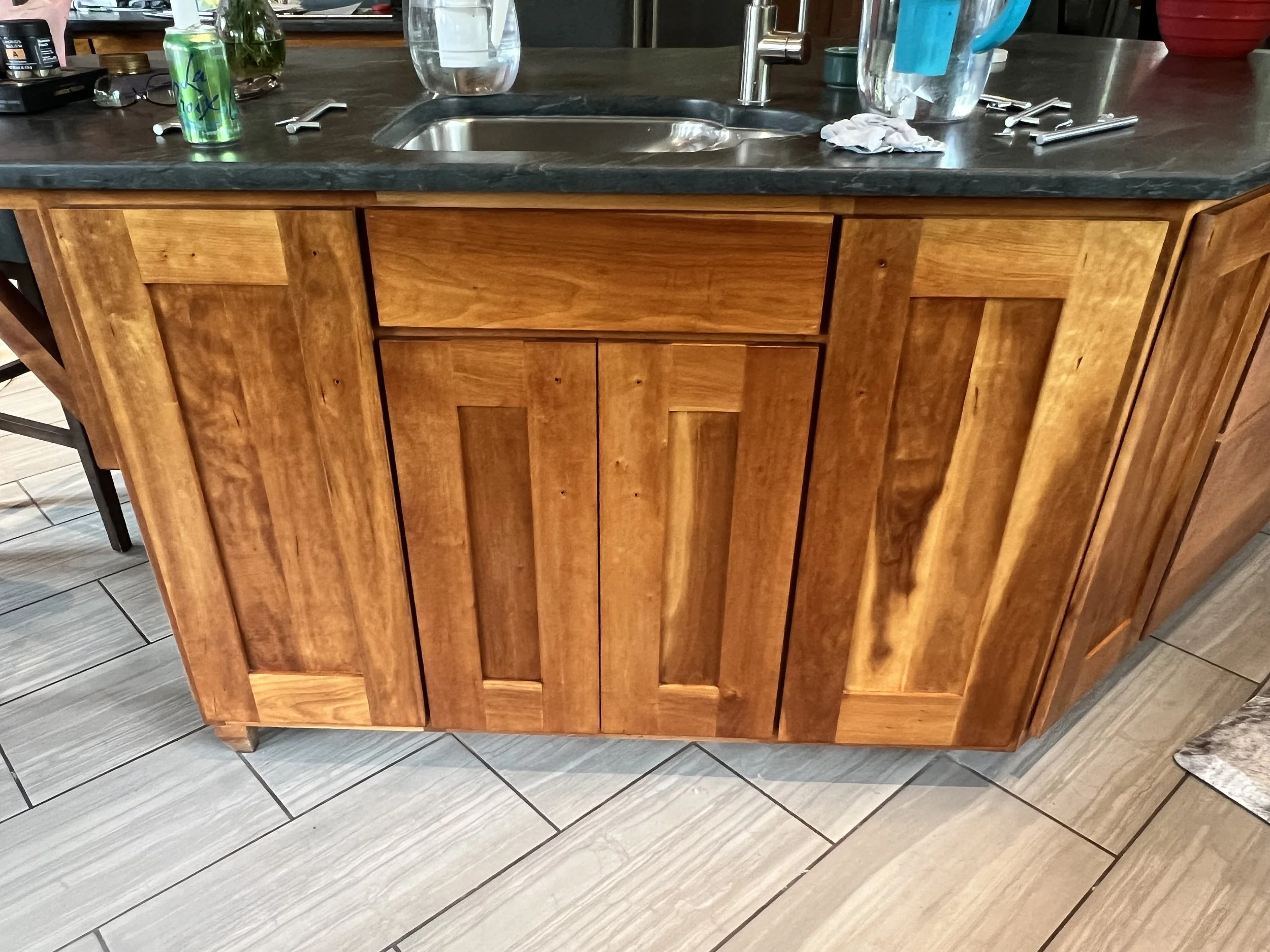 A kitchen counter with a wooden cabinet below, a stainless steel sink, and various items on the counter including a bottle of water, a glass, and some utensils.