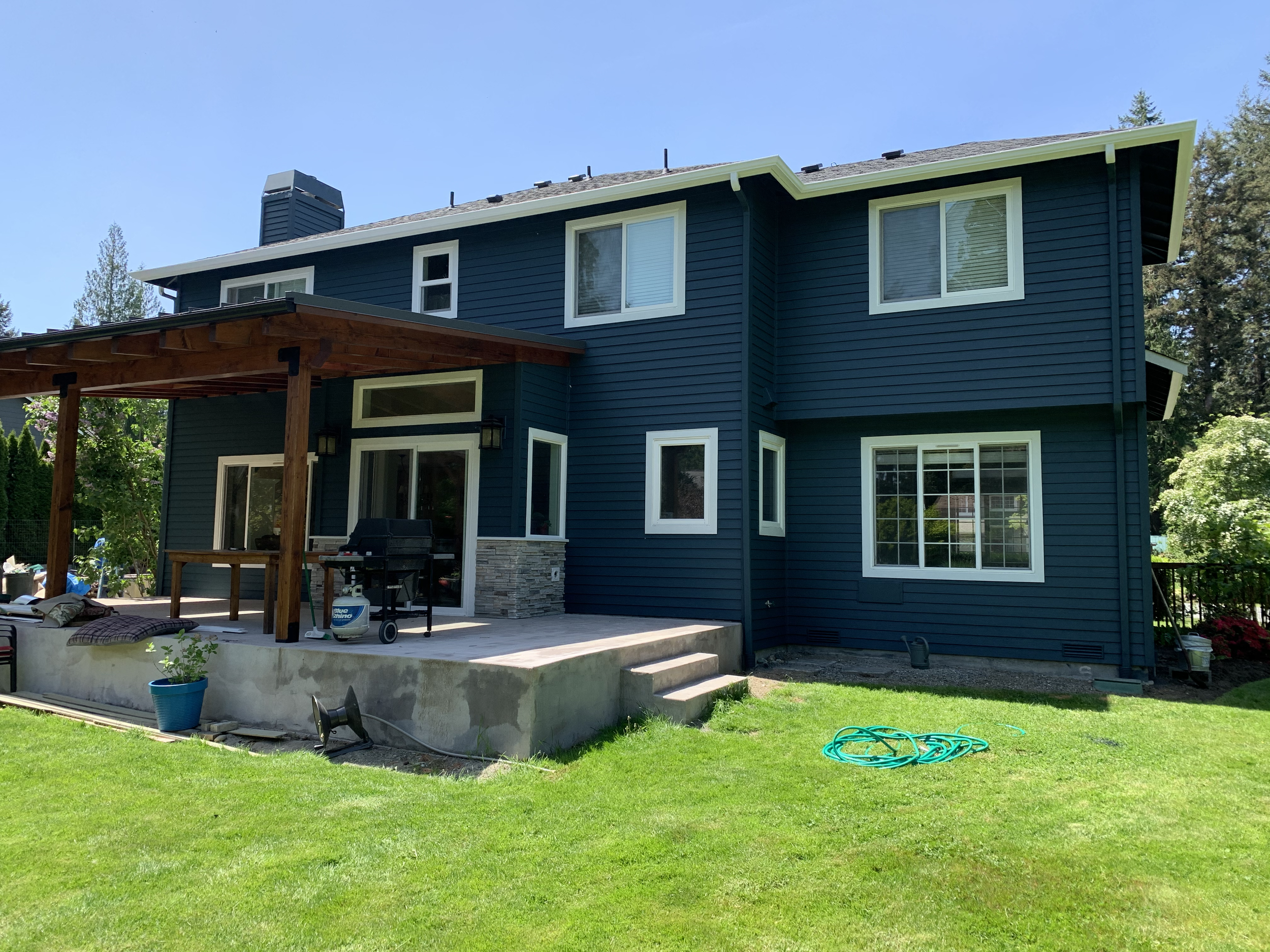 A blue two-story house with white window frames and a partially covered concrete patio in the backyard. There is a wooden pergola, a grill, and a blue pot with a plant on the patio. The yard has green grass, a garden hose, and trees.