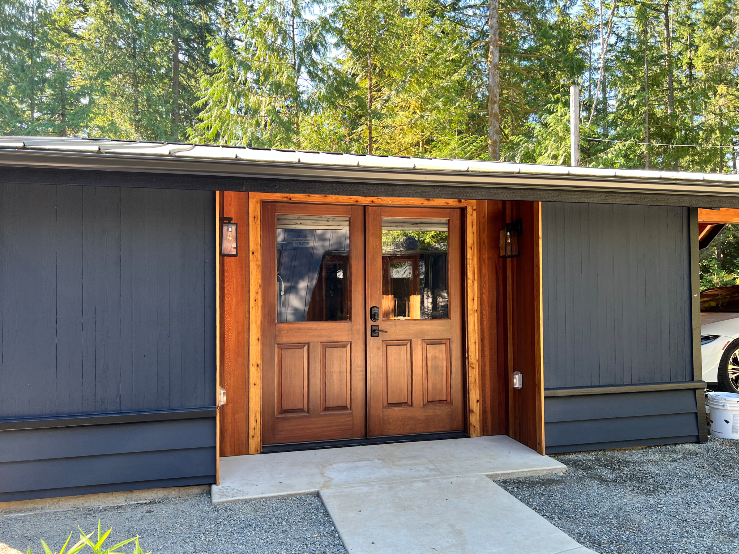 Front view of a modern house with a wooden door, black exterior walls, and exterior lighting, surrounded by trees.