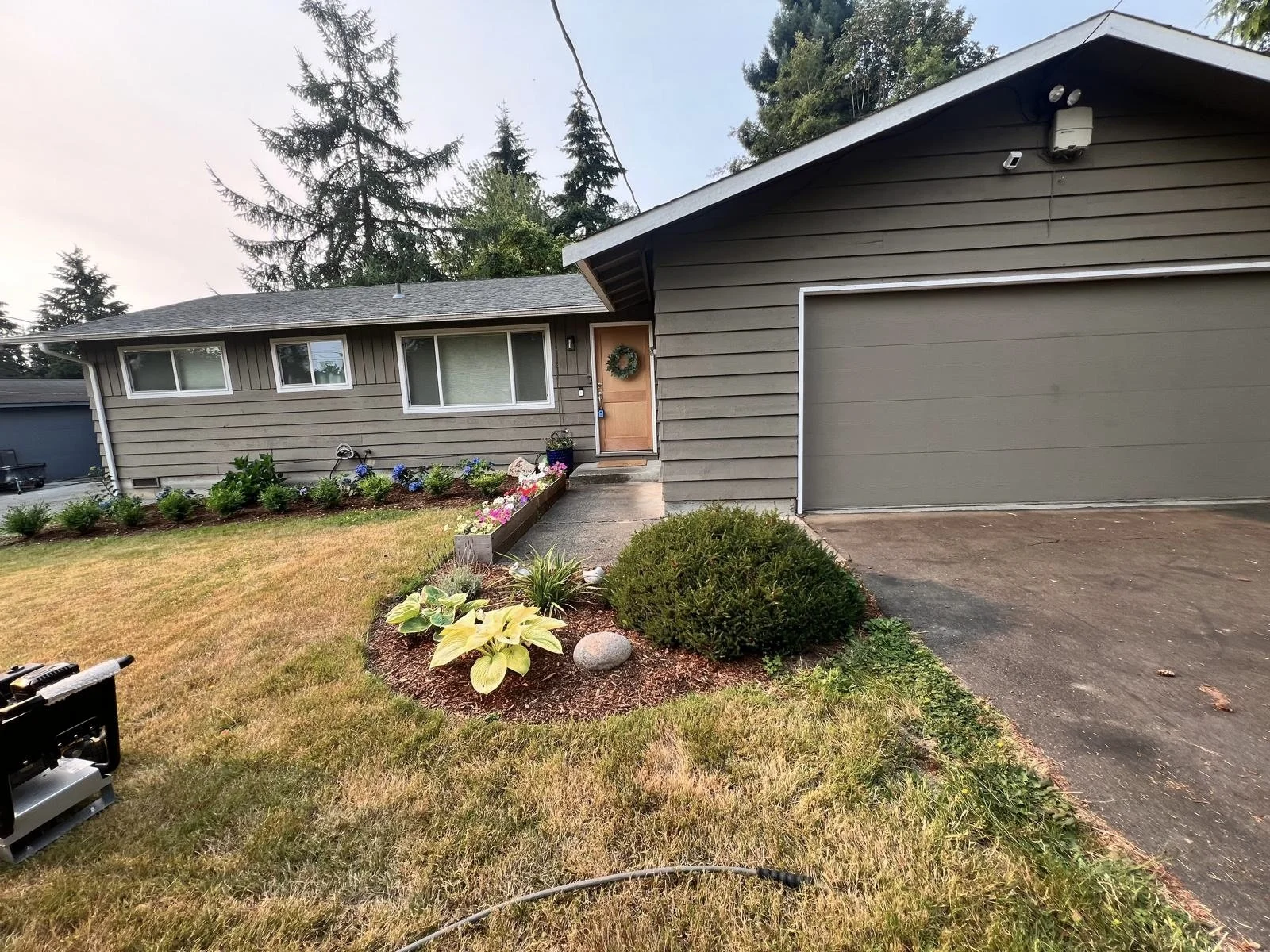 A single-story house with gray horizontal siding, a brown front door with a wreath, and a two-car garage with gray doors, surrounded by a well-maintained lawn, flowerbeds with various plants and flowers, and tall trees in the background.