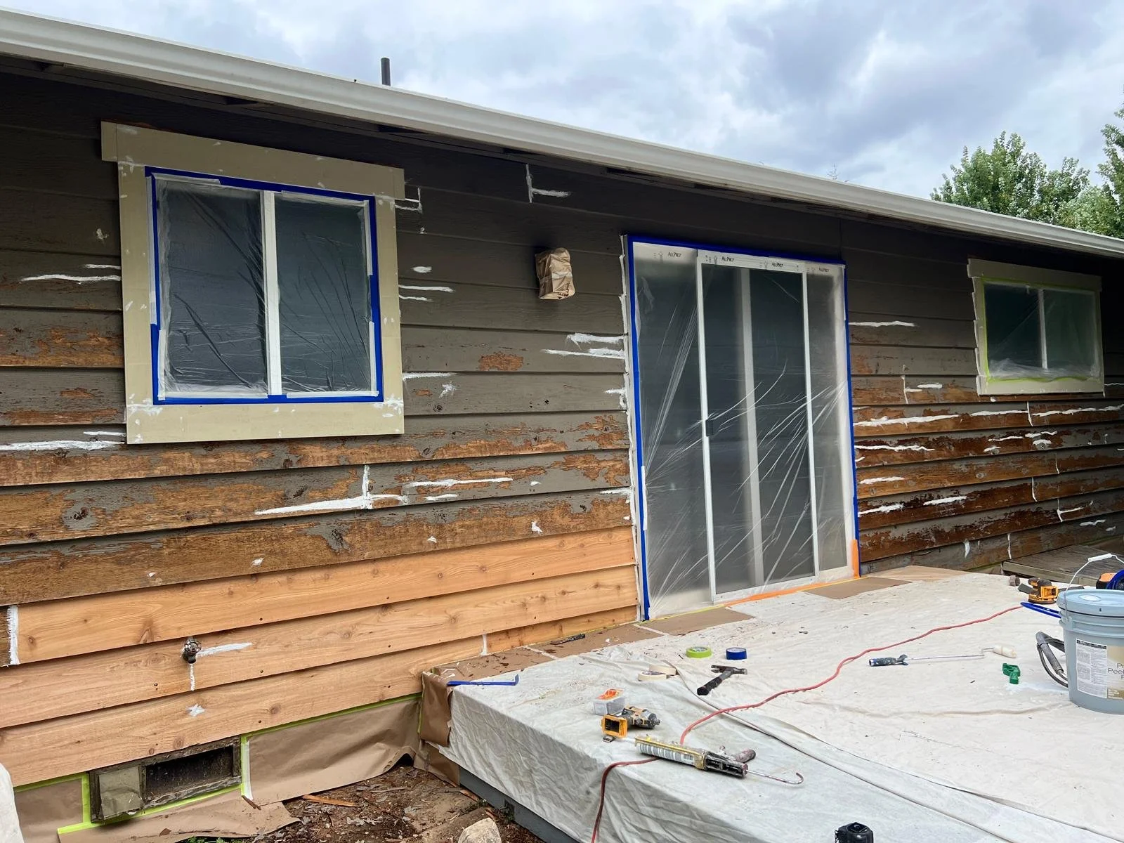 Exterior of a house under renovation, with a wooden siding partially removed, covered windows, construction tools, and materials on a covered porch.
