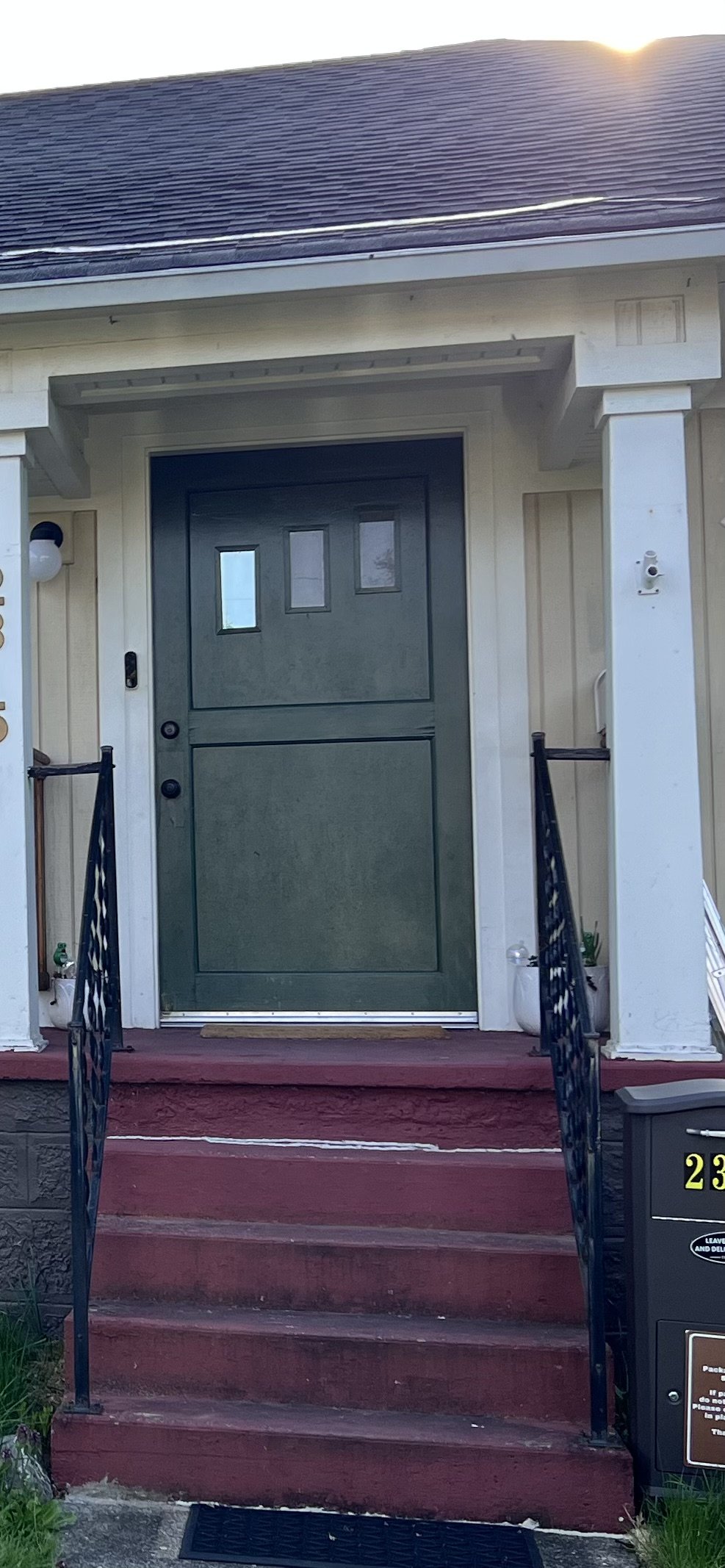 Green front door with, flanked by a grey railing on stairs, outside of a house with cream siding.