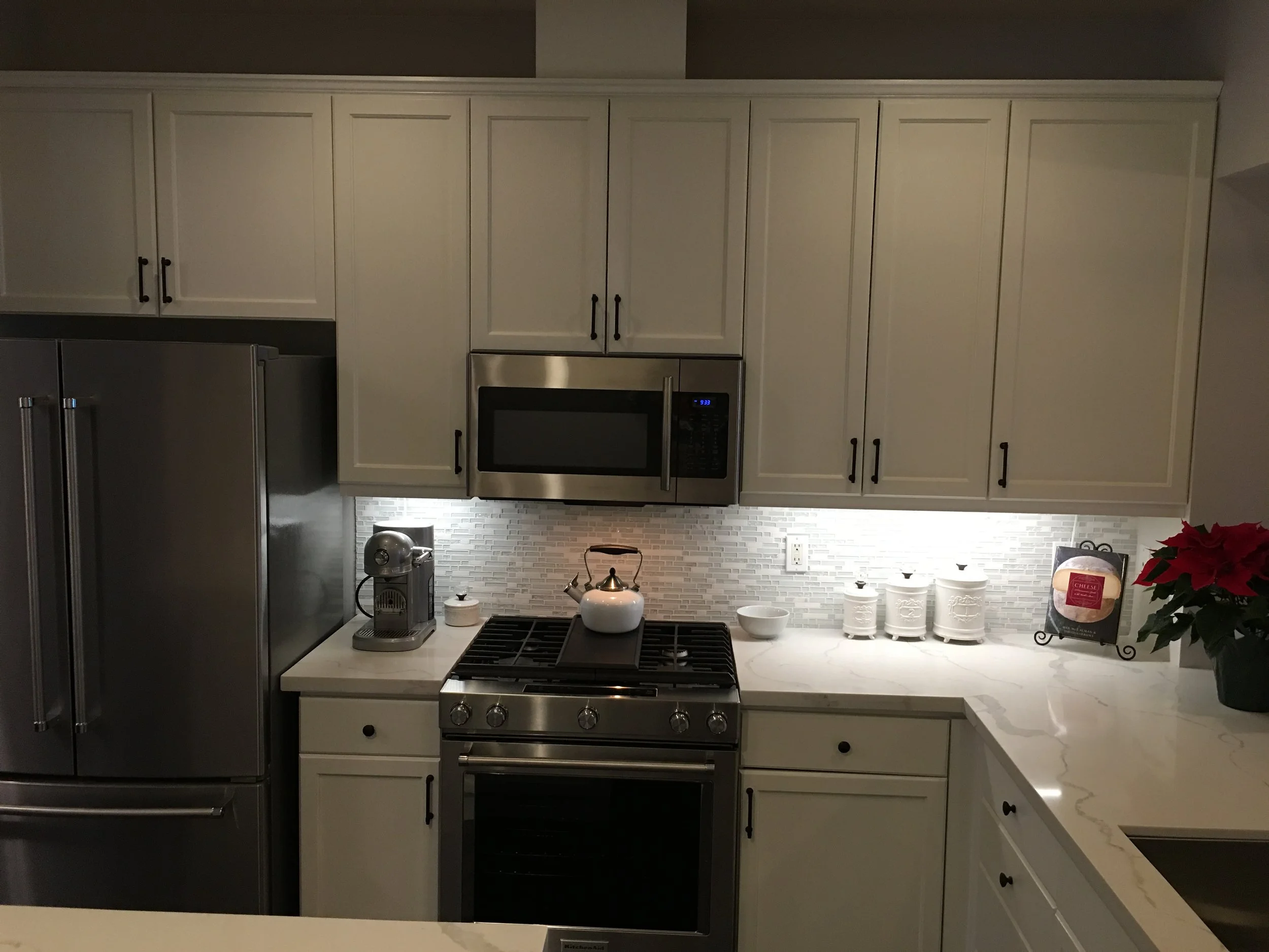 A modern kitchen with cream-colored cabinets, stainless steel refrigerator, microwave, and oven. The counter has a white teapot, a coffee machine, and decorative containers, with holiday-themed poinsettia plant on the right.