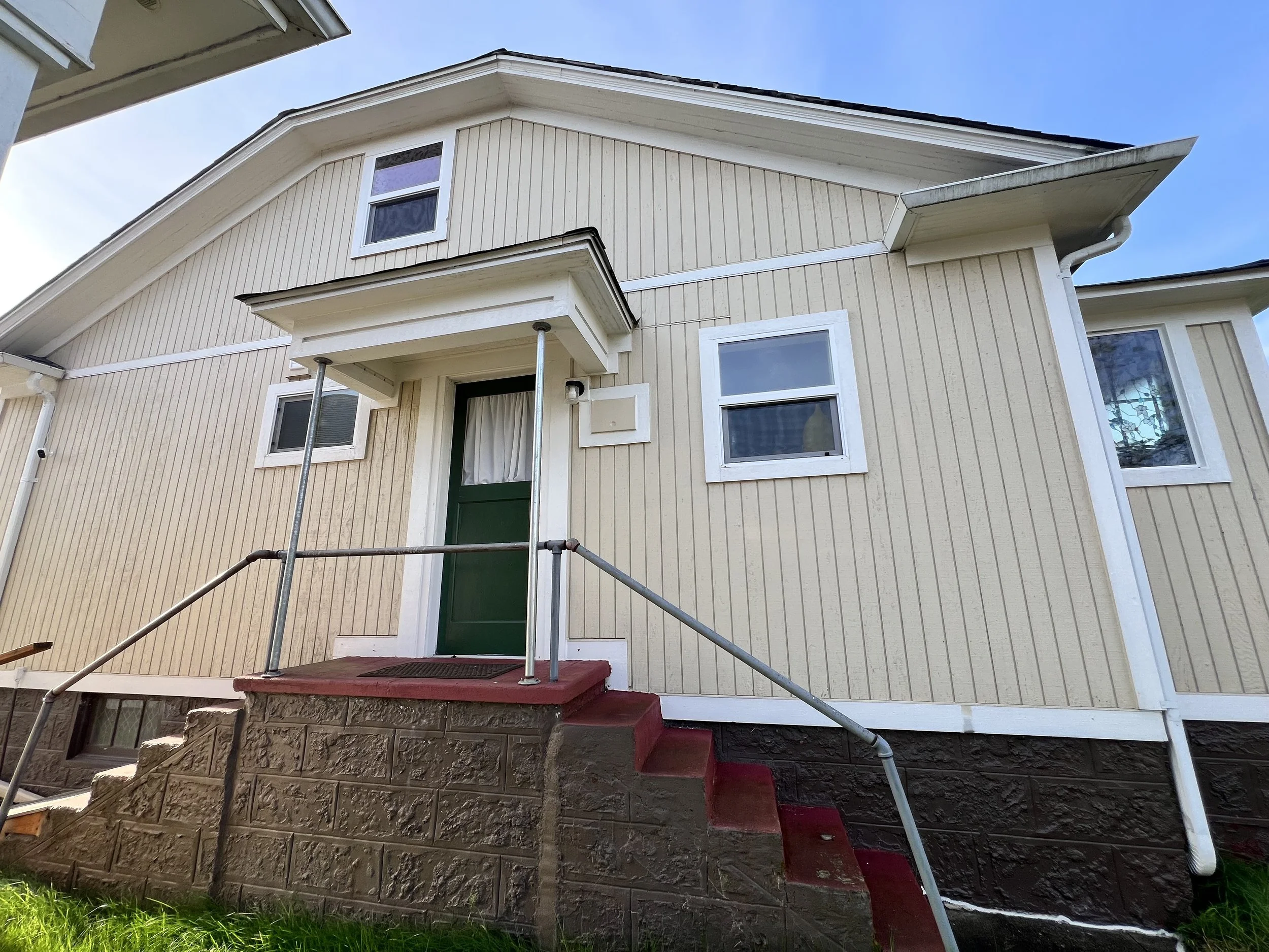 Front of a beige wooden house with white trim, green front door, and red steps.