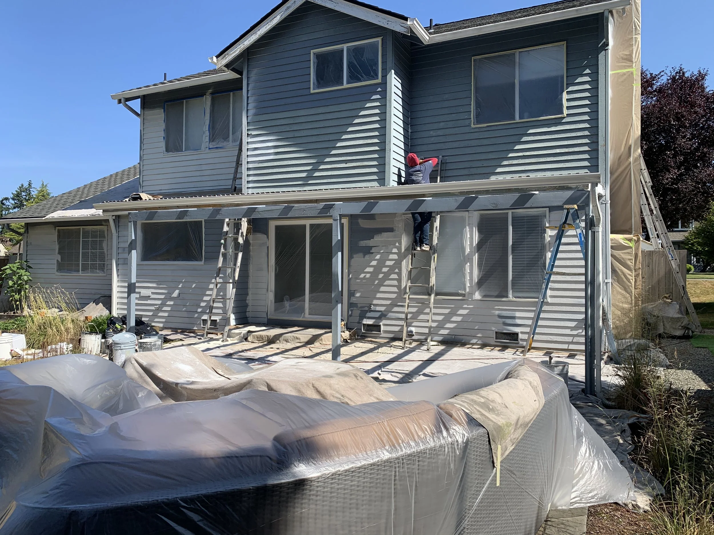 Two people remodeling the exterior of a blue two-story house, working on constructing a new screened porch or extension, using ladders and scaffolding, with construction materials and tarps on the ground.